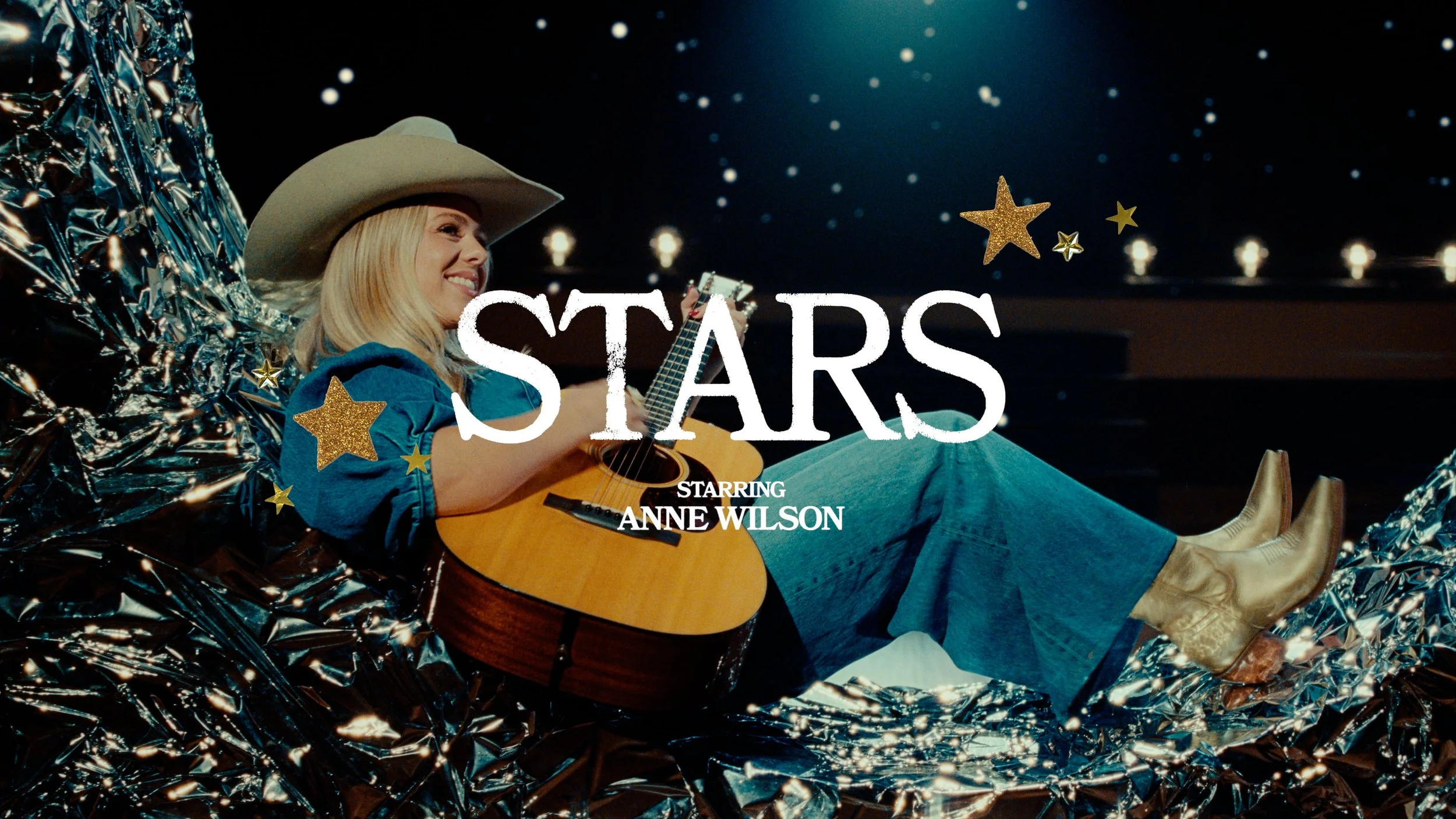 A woman lying on a shiny metallic surface, wearing cowboy boots, a blue shirt, and a cowboy hat, playing an acoustic guitar under starry lights.