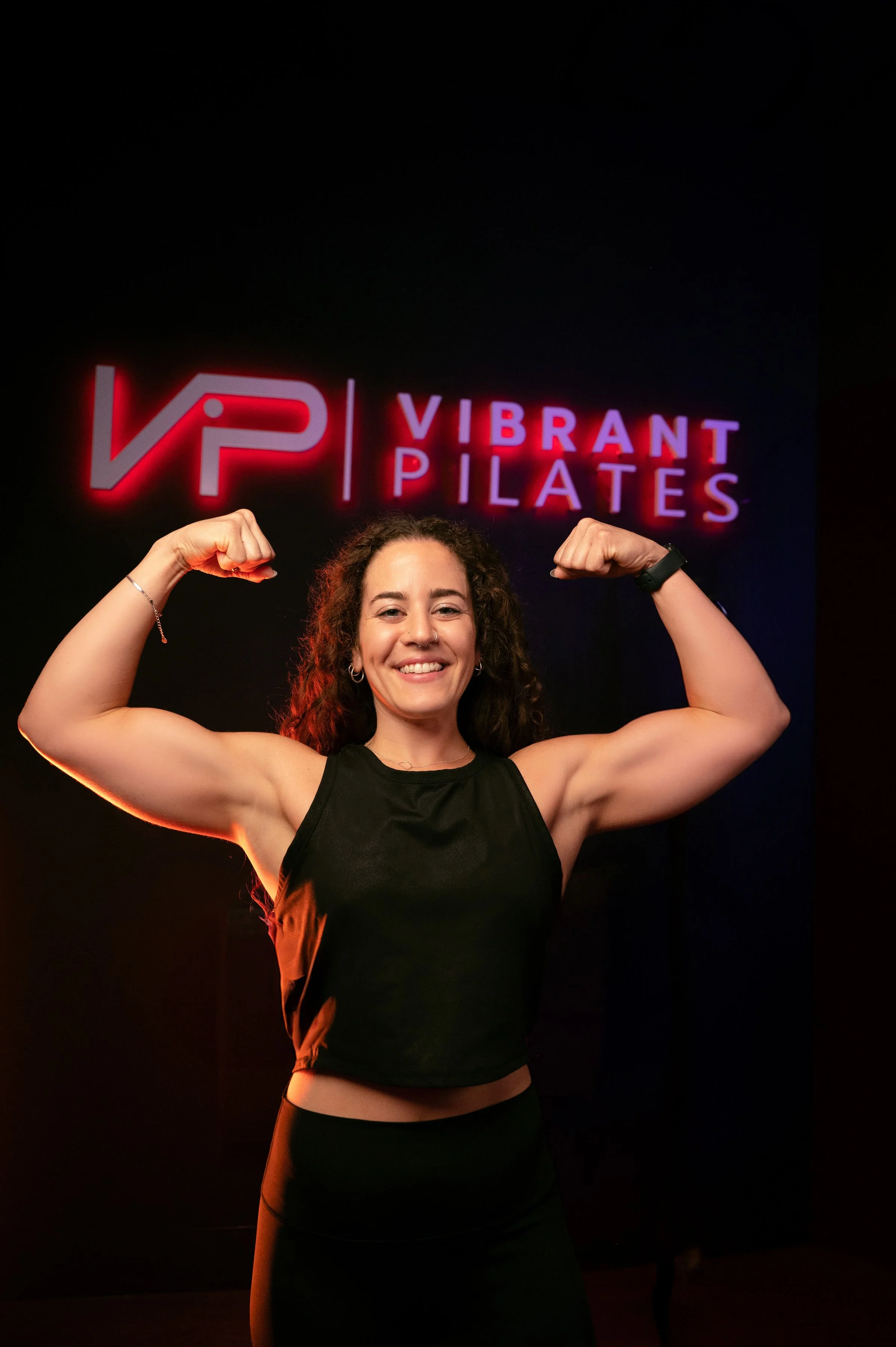 A woman flexing her biceps in front of a neon sign that reads 'Vibrant Pilates' at Vibrant Pilates studio.