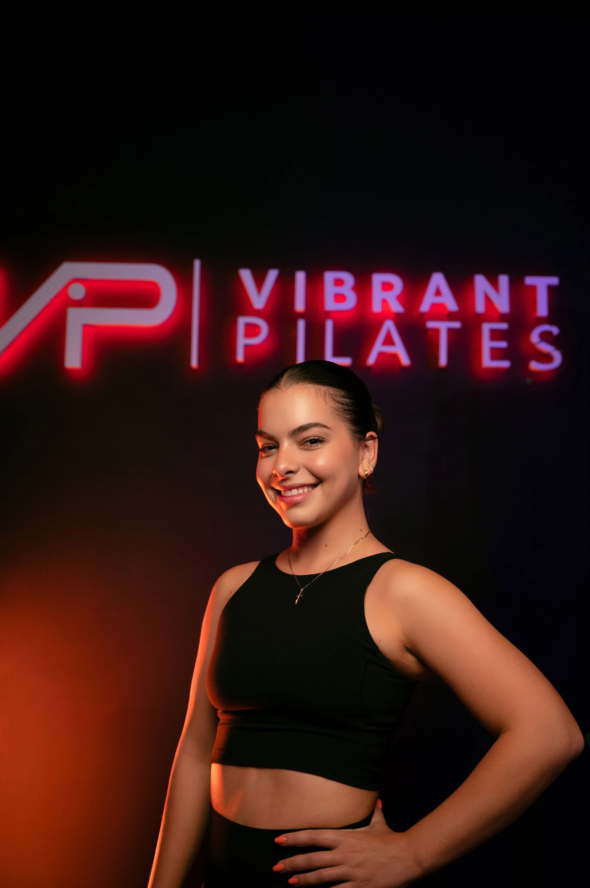 A young woman in workout attire standing in front of a neon sign that reads 'VIBRANT PILATES' and a logo.