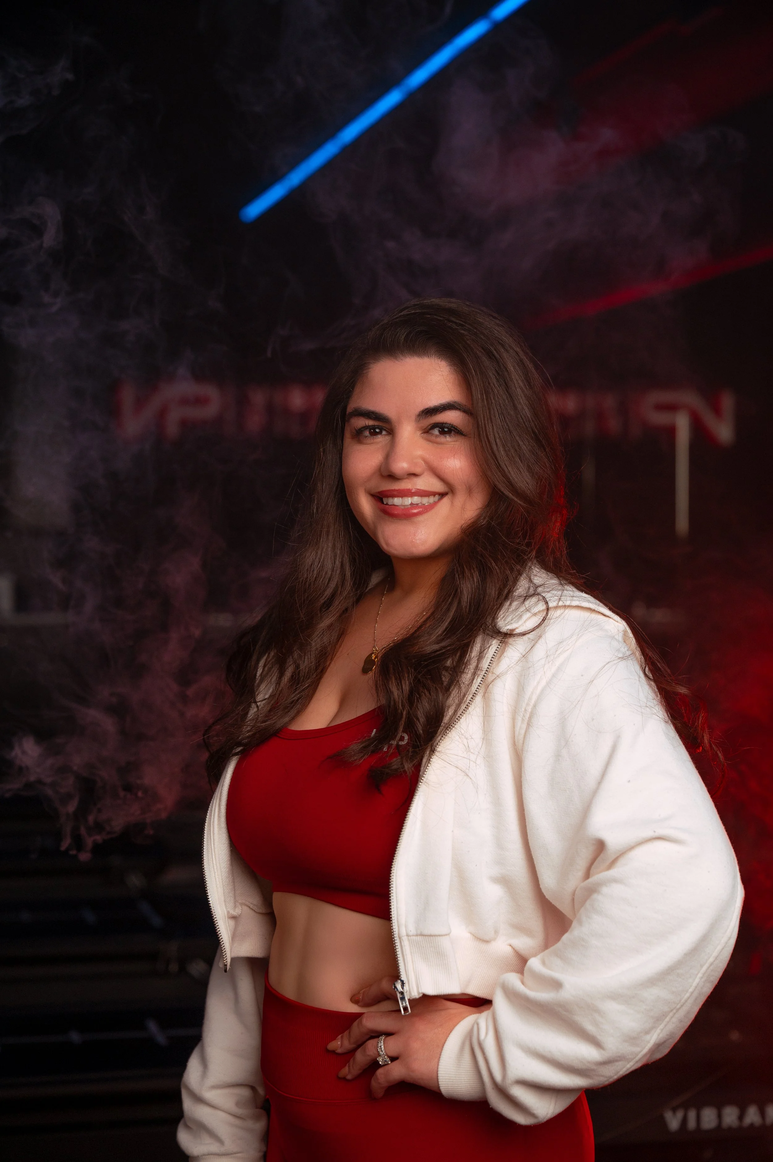 A woman with dark curly hair wearing a white baseball cap, black sleeveless top, and a gold necklace, smiling in front of a black wall with red illuminated letters spelling 'HYBRID PILATES'.