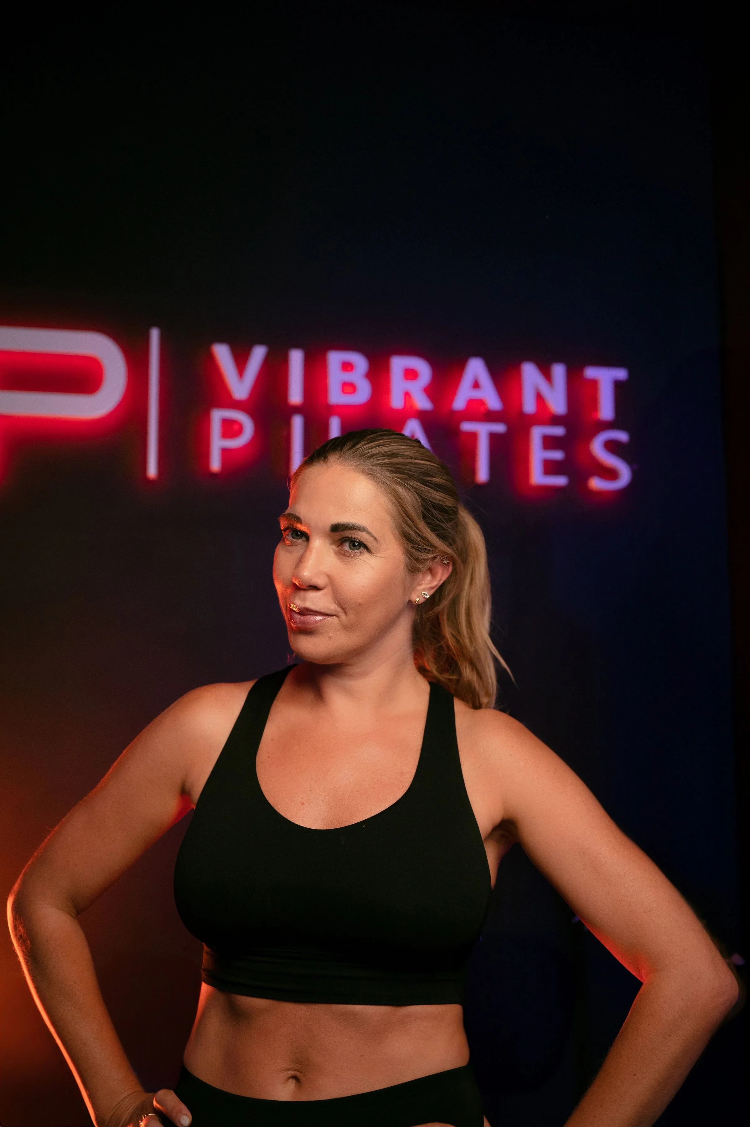 A woman with blonde hair in a ponytail, wearing a black sports bra, stands in front of a dark background with neon signs reading 'VIBRANT PIRATES'.