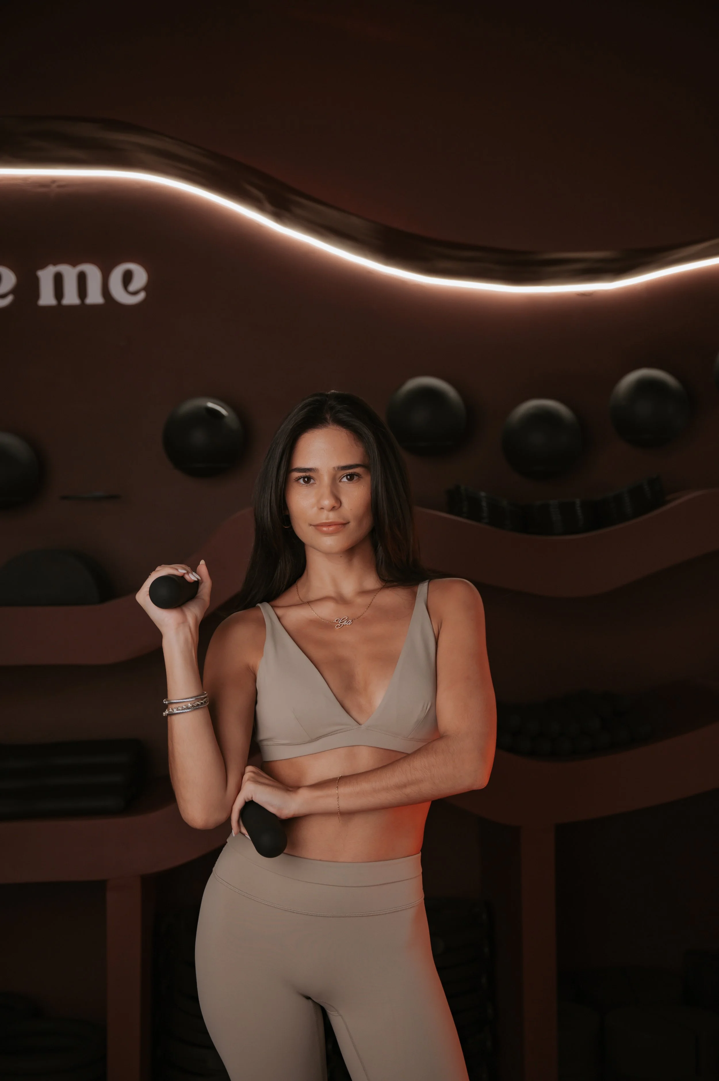 A young woman with long dark wavy hair standing in front of a black background with a neon sign that reads 'Vibrant Plates' and a logo with the letters 'VP'. She is wearing a black sleeveless top, a gold necklace, and has her arms crossed.