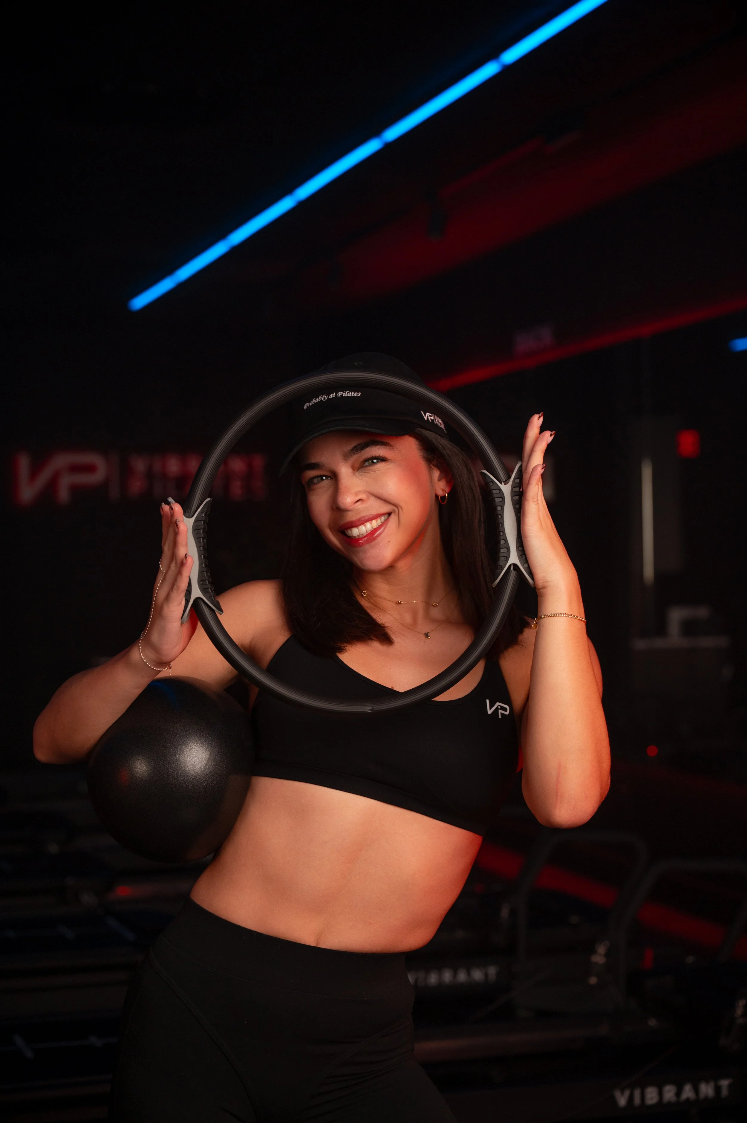 A young woman standing with arms crossed in front of a sign that reads 'Vibrant Pilates' with a stylized 'VP' logo, in a dark room with red and purple lighting.