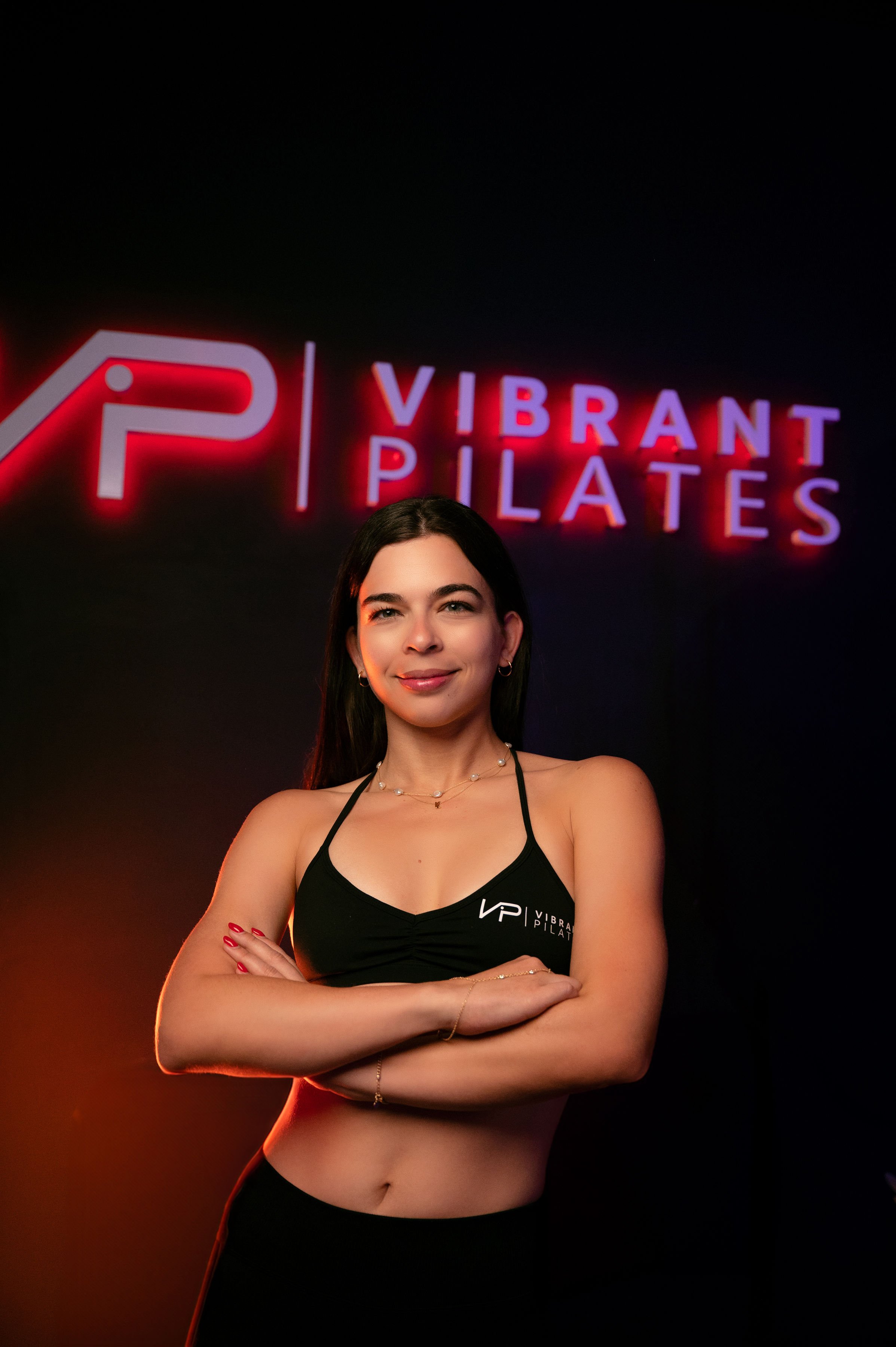 A young woman standing with arms crossed in front of a sign that reads 'Vibrant Pilates' with a stylized 'VP' logo, in a dark room with red and purple lighting.