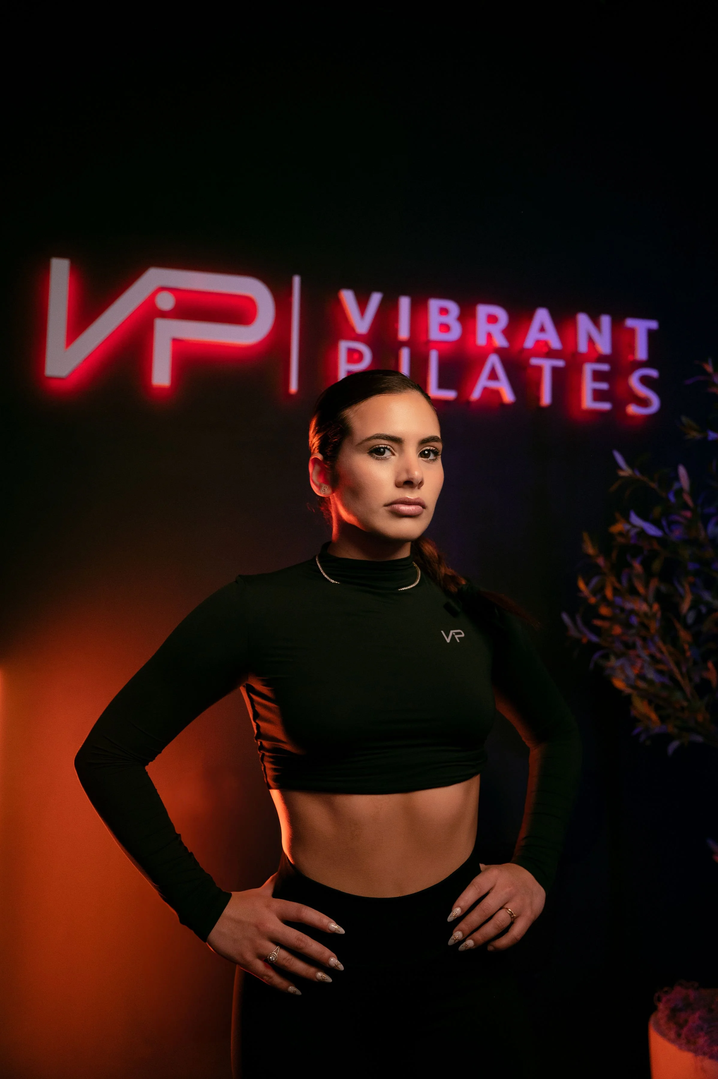 A woman with dark hair in a braid, wearing a black athletic crop top with 'VP' on it, standing indoors in front of a neon sign that reads 'Vibrant Pilates' and 'VP', with both hands on her hips.