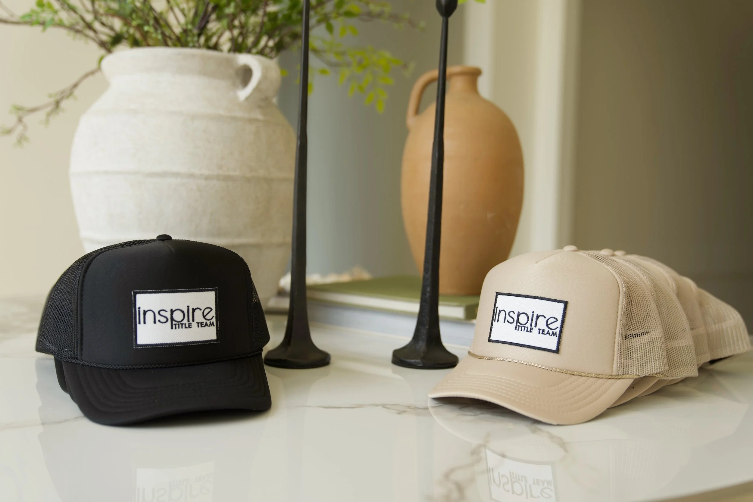Black and beige mesh caps with 'inspire LITTLE TEAM' patches, displayed on a white marble surface with decorative vases and books in the background.