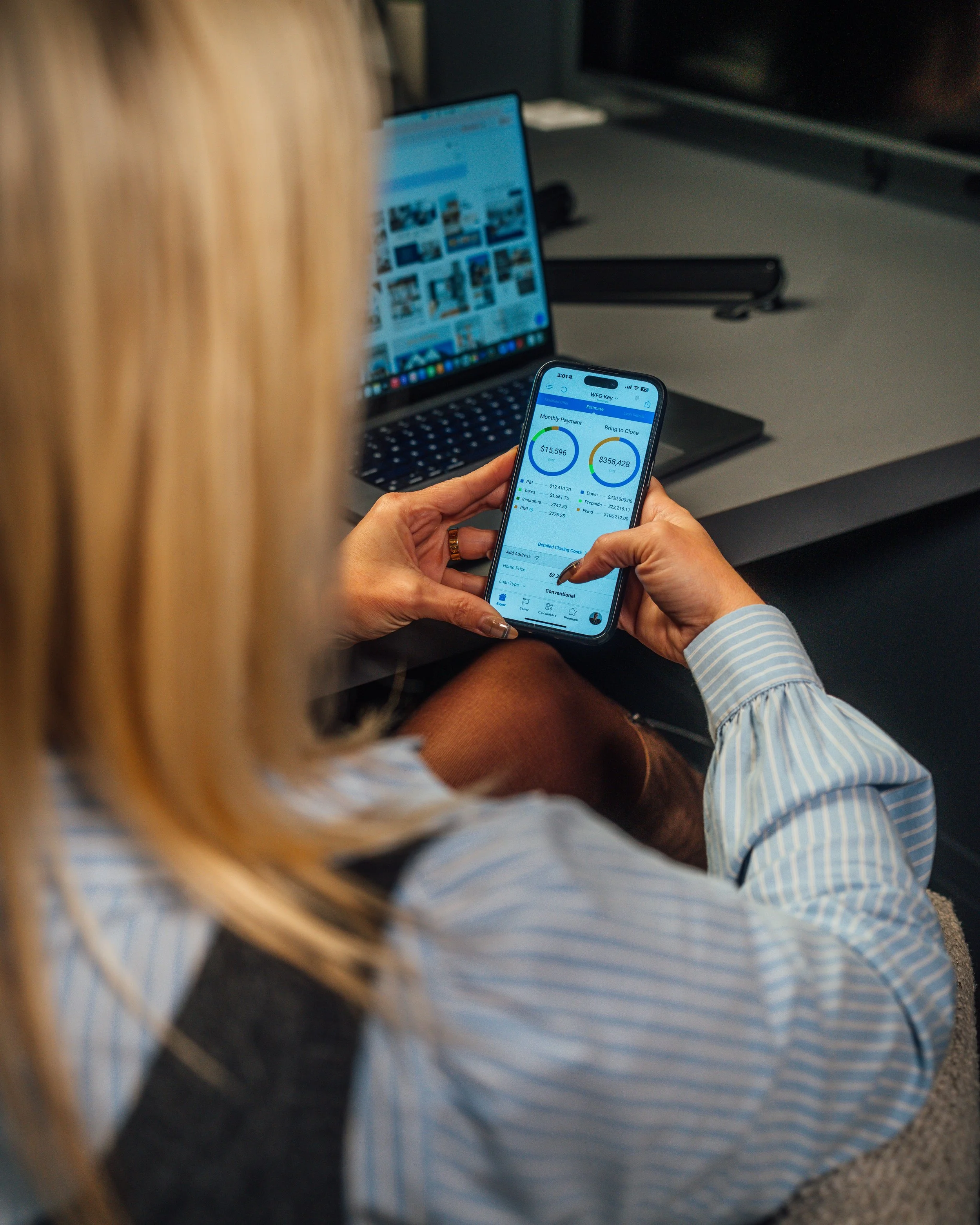 A person with blonde hair is looking at a smartphone displaying a financial app, with a laptop in the background on a desk.