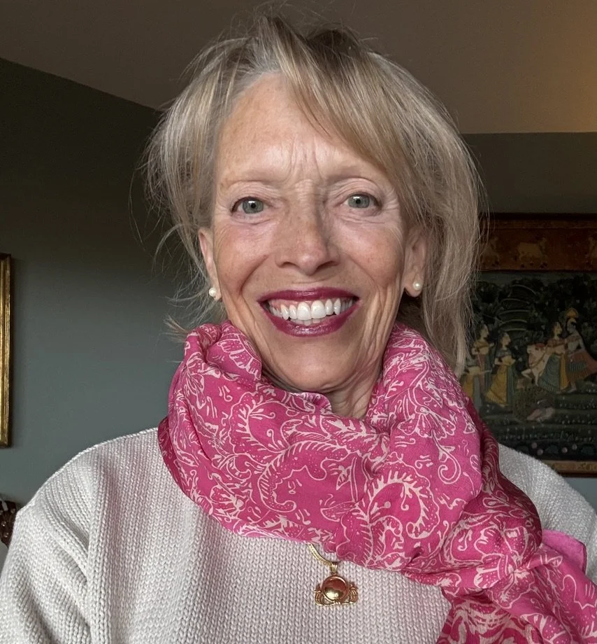 A smiling woman with blonde hair, wearing white pearl earrings, a pink patterned scarf, and a white sweater, standing indoors with artwork in the background.