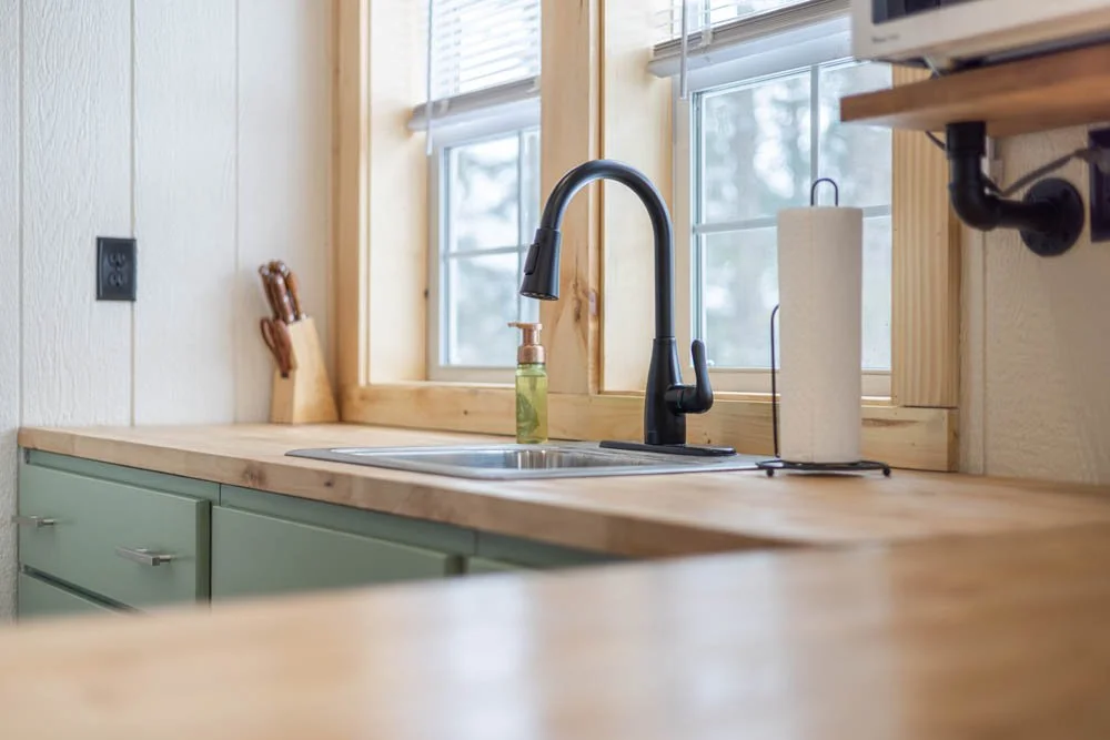 Cozy kitchen interior with wooden countertops, black faucet, kitchen sink, paper towel holder, knife block, and green lower cabinets, set against a window with natural light.
