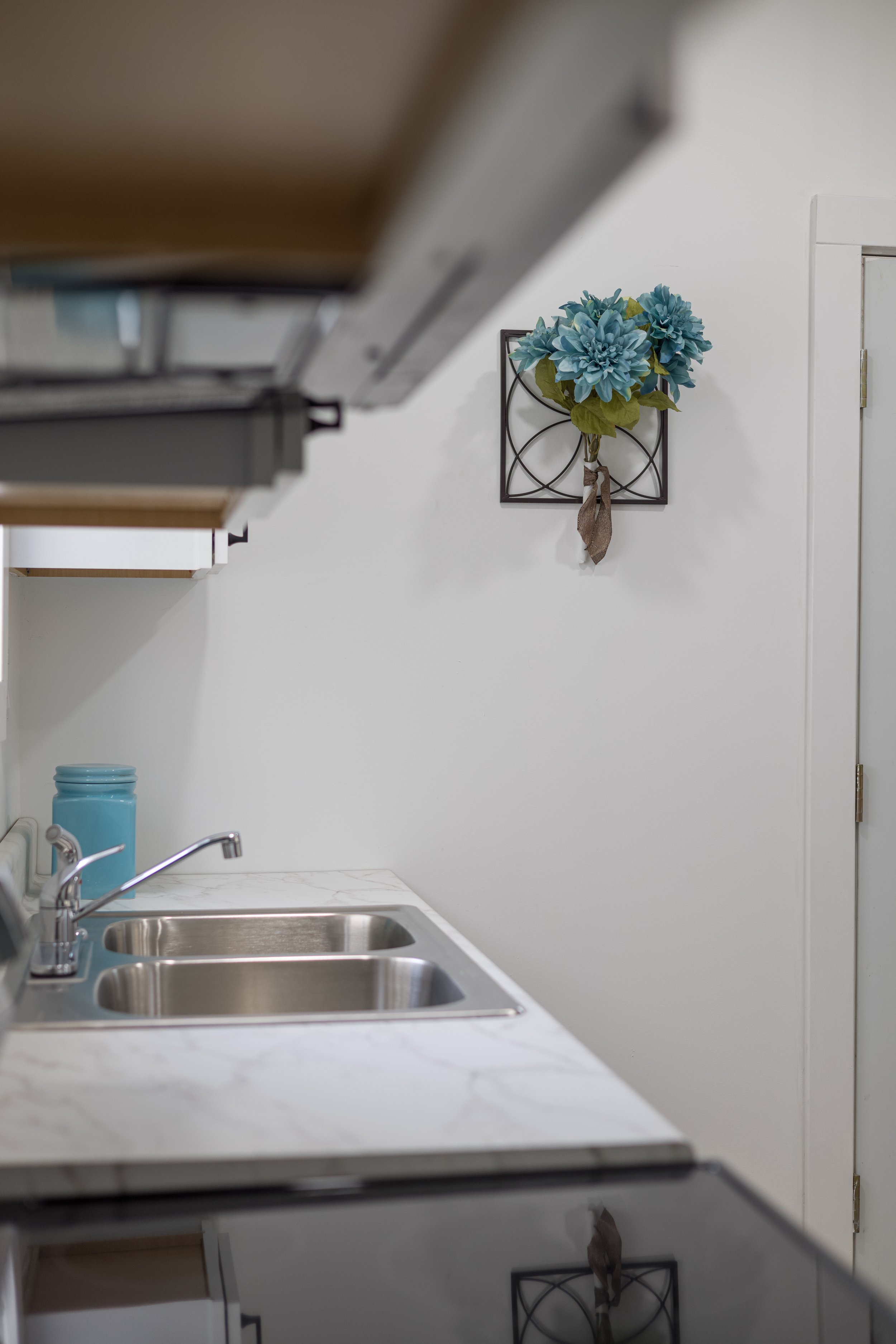 Modern kitchen interior with stainless steel sink, white countertop, turquoise canister, and wall-mounted blue floral decoration.