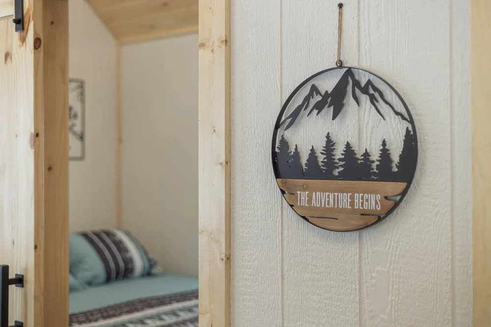 Interior of a wooden cabin with a decorative wall sign featuring a mountain and forest silhouette, and the phrase "The Adventure Begins." A bed with striped bedding is visible inside the room.