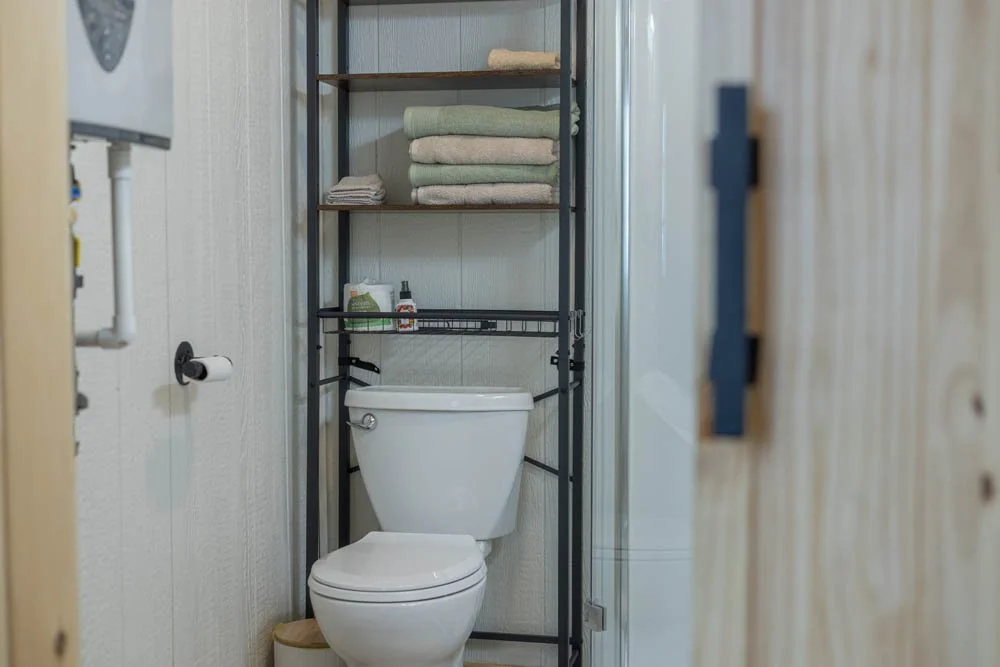 Small bathroom with a toilet, over-the-toilet shelving unit, towels, and toiletries, next to a light-colored wooden door.