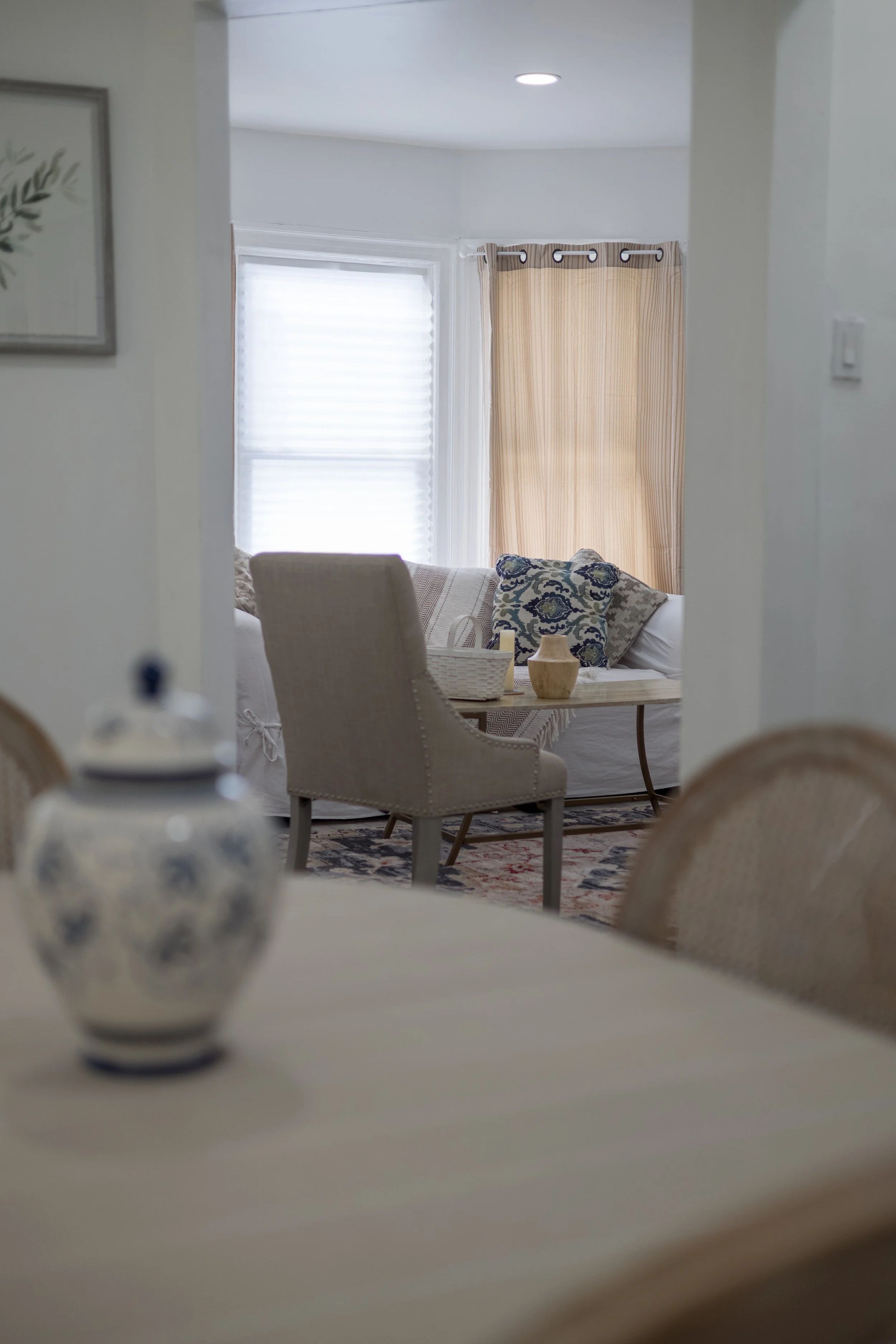 Cozy living room with a beige chair, patterned cushions, a small table with a vase, and a partial view of a dining table with a decorative jar.