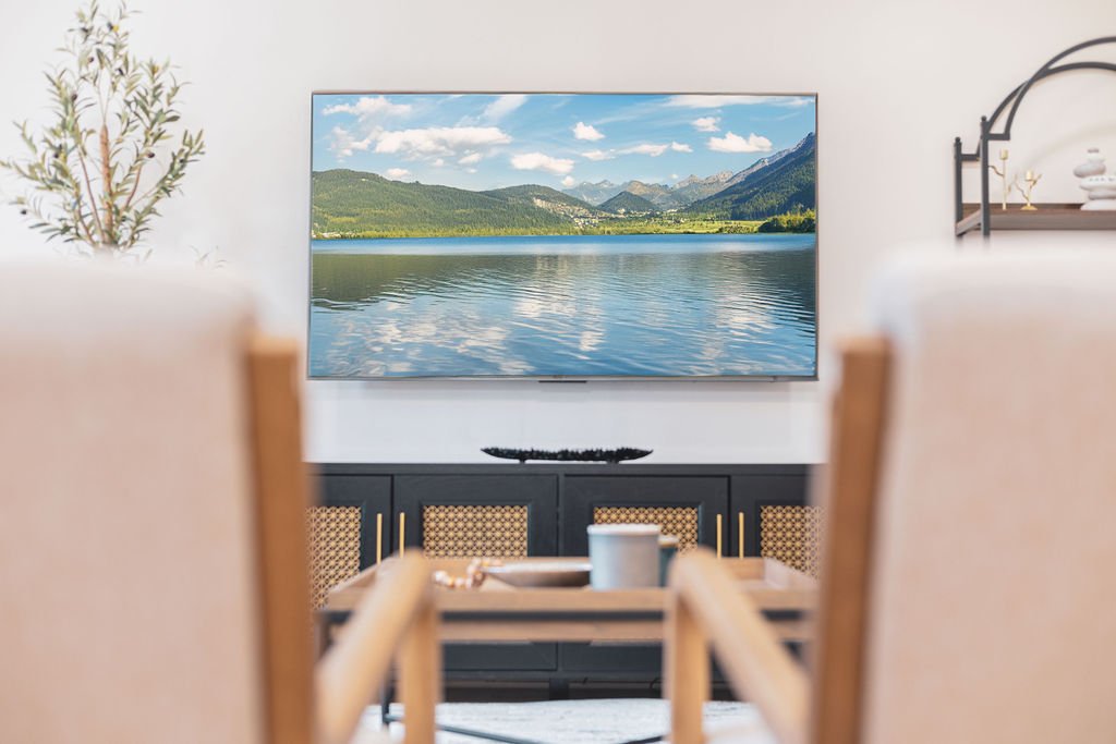 Living room setup with a wall-mounted TV displaying a scenic lake and mountain view, chairs in the foreground, and modern decor.