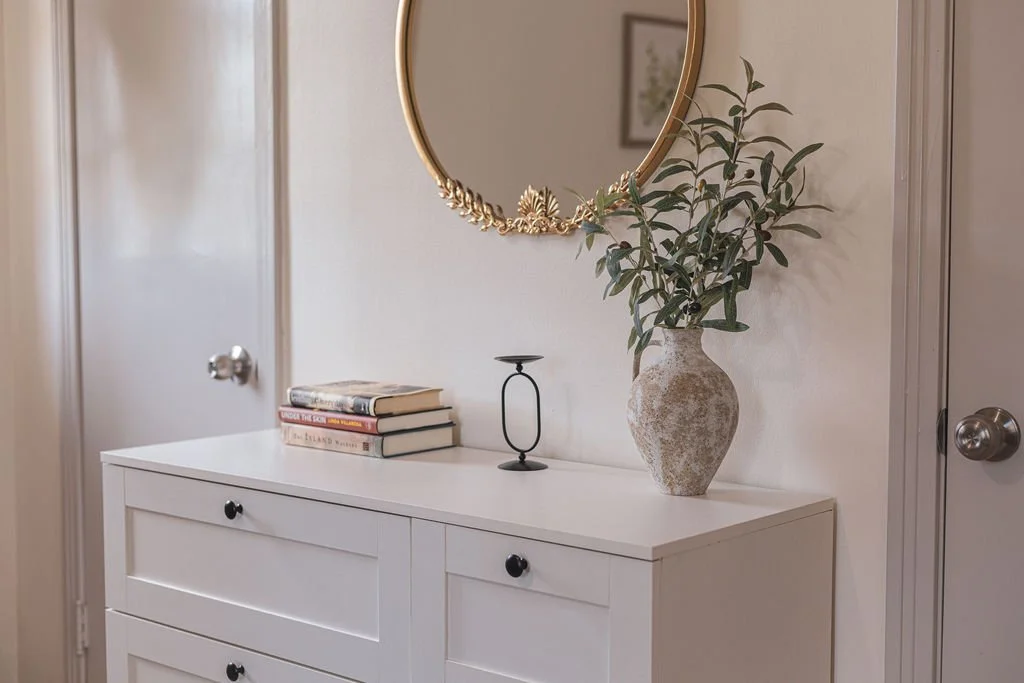 White dresser with books, candle holder, vase with greenery, and a round gold mirror above.