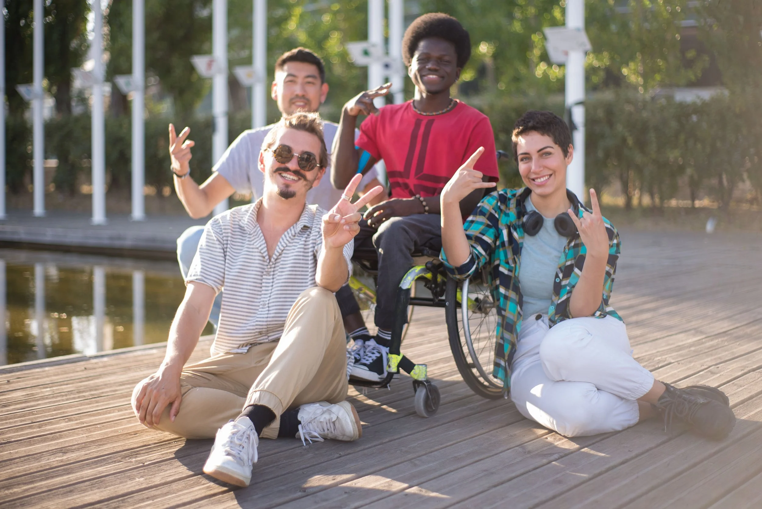 Diverse group of four friends, one in a wheelchair, smiling and displaying various hand-gestures of peace and joy.