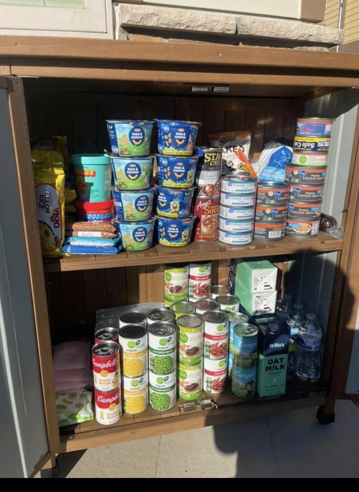 Inside an open outdoor food pantry showing shelves stocked with canned soups, instant noodles, pasta cups, bottled water, toilet paper, wipes, and hygiene items.