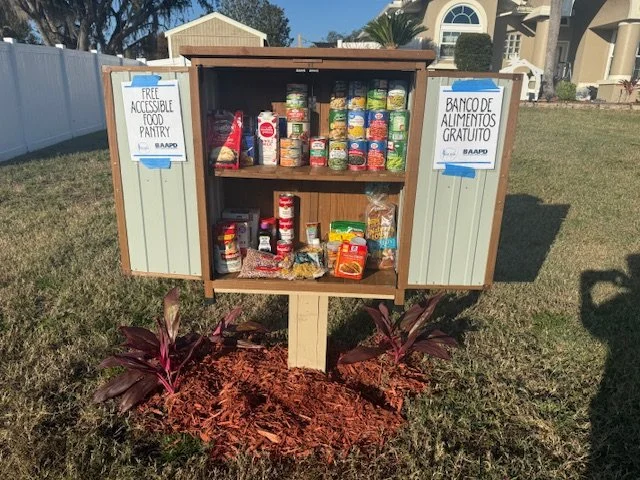 Outdoor free food pantry mounted on a post, doors open to reveal shelves of canned goods and dry food items.