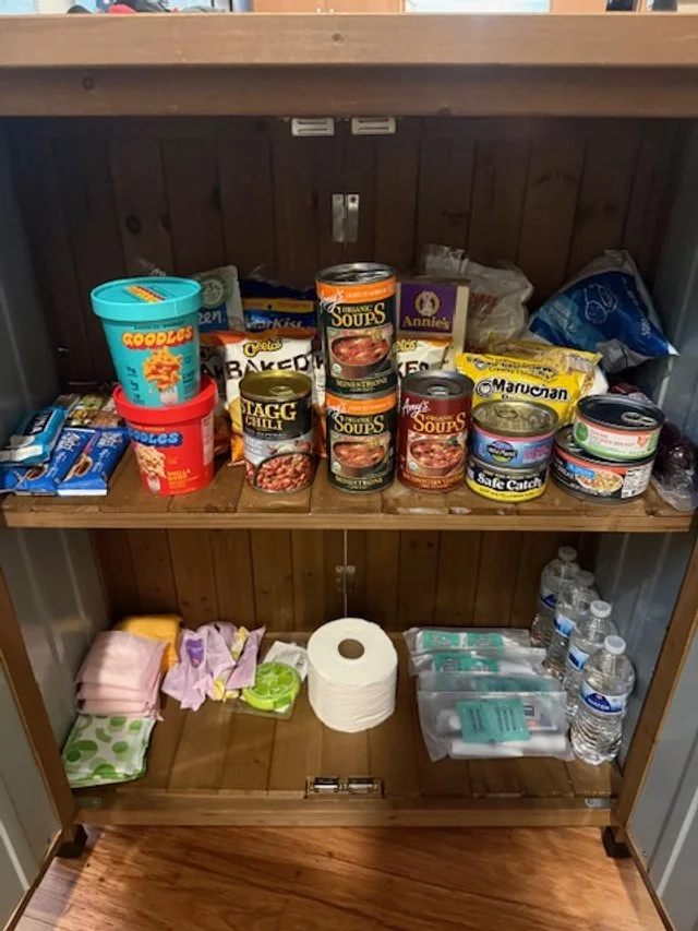 Top-down view of an open outdoor pantry stocked with bottled water, canned vegetables, boxed meals, and packaged foods.