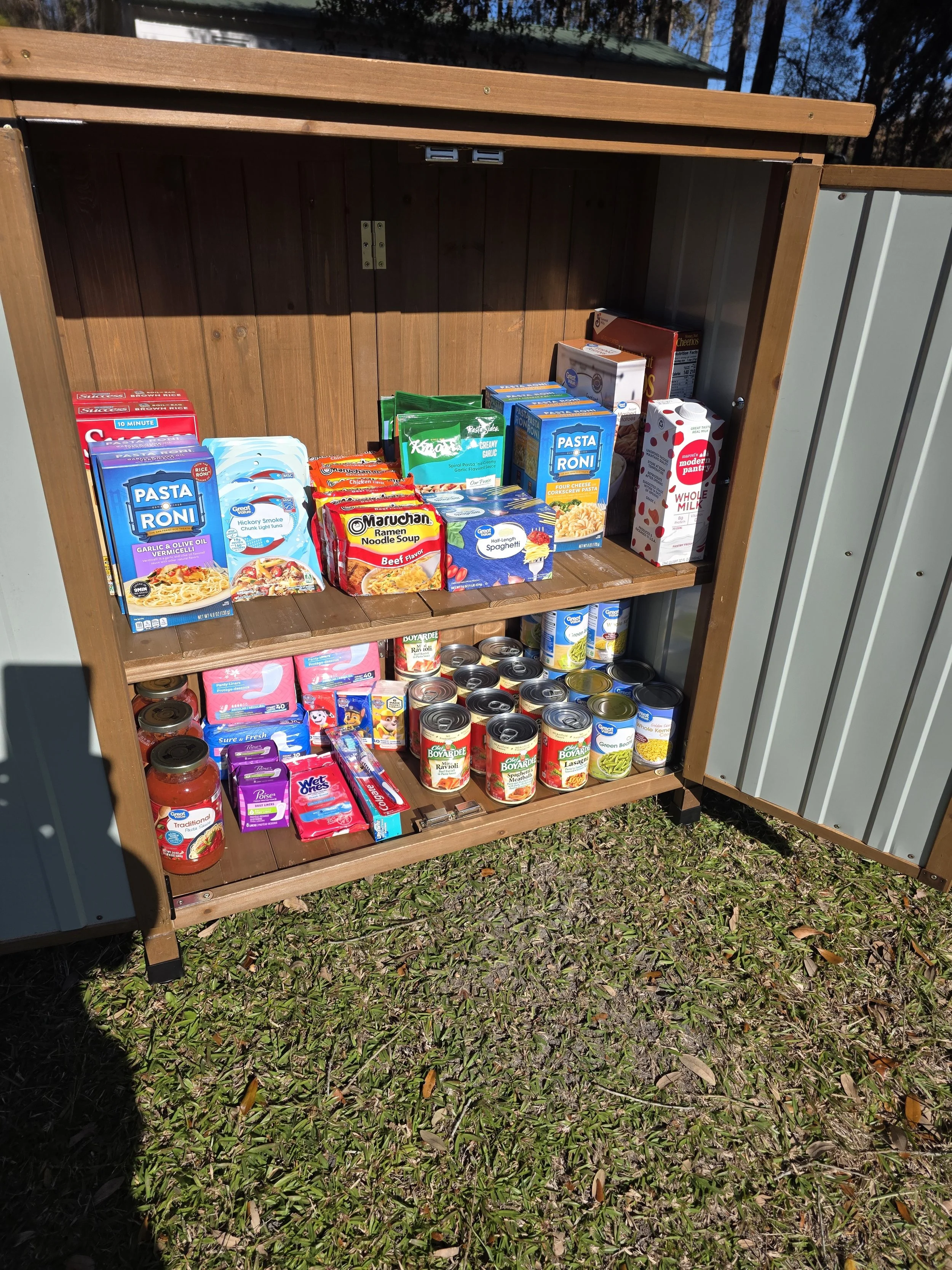 Another open outdoor pantry filled with macaroni cups, canned soup, canned vegetables, oatmeal, water bottles, and oat milk.