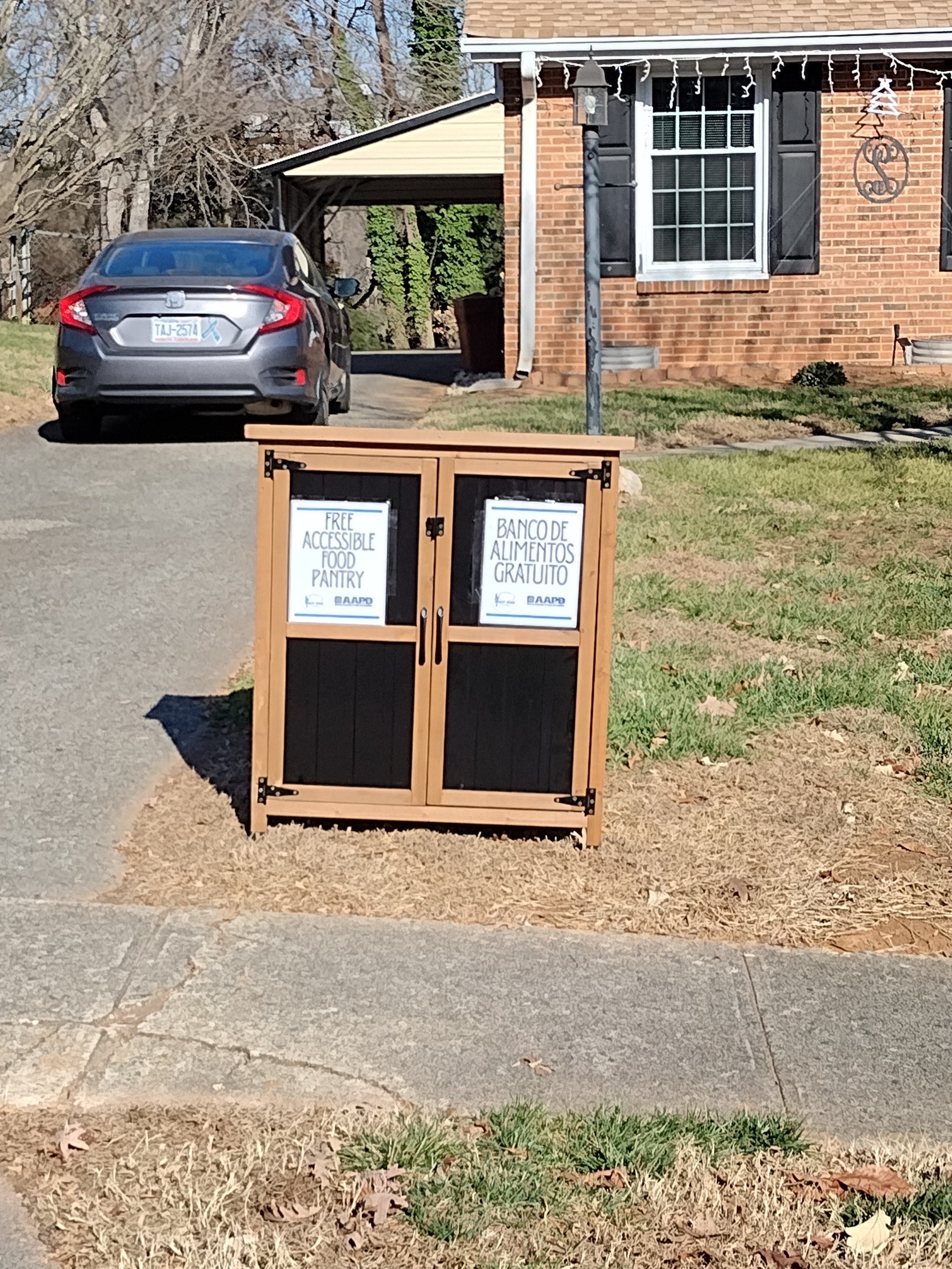 Photo of a food pantry that is closed and has signs on the outside that say "Accessible Little Free Pantry" in Spanish and English. This pantry sits next to the sidewalk in front of a brick house or building.