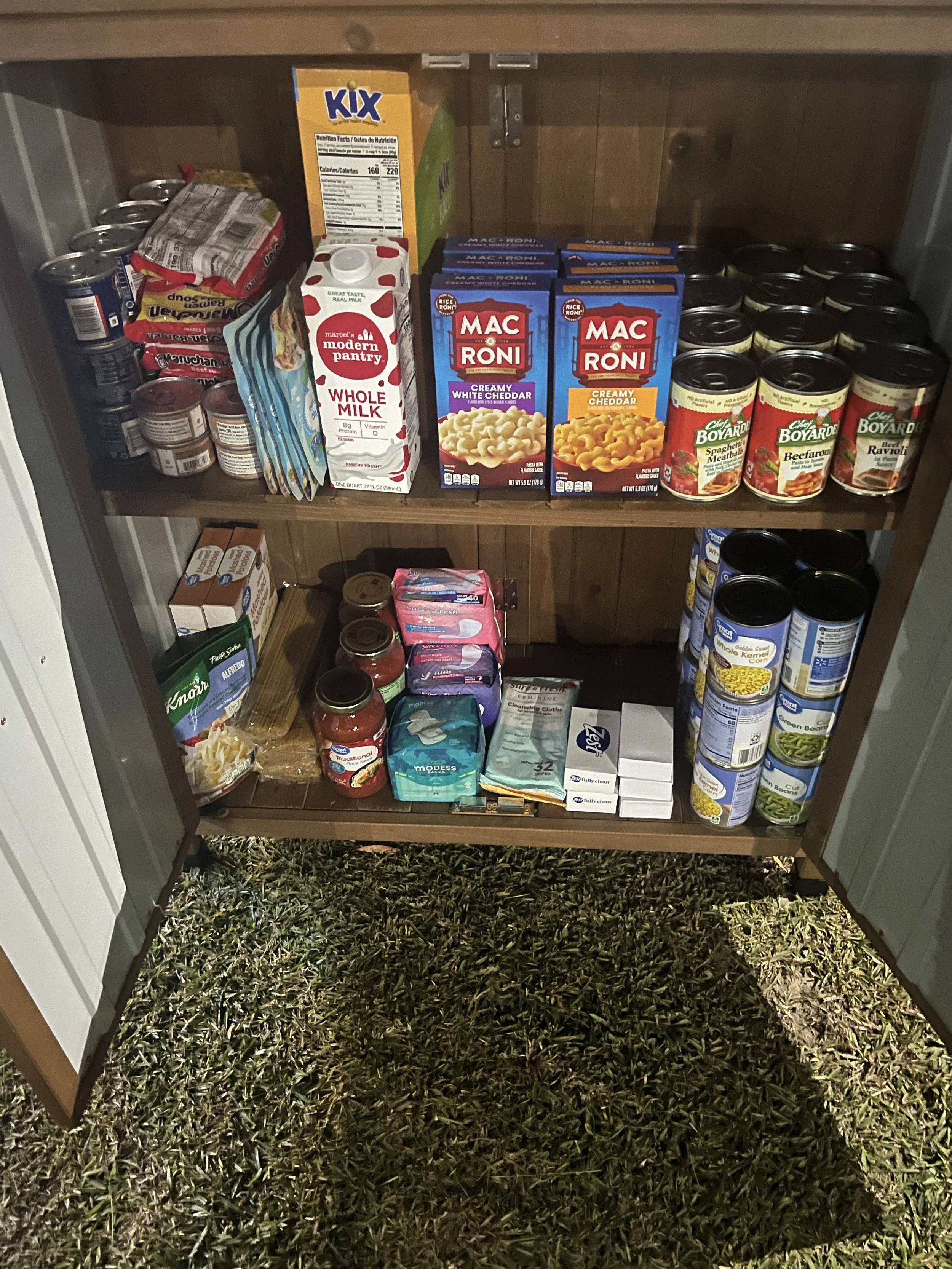 Inside an open outdoor food pantry showing shelves stocked with boxed meals, canned foods, pasta, whole milk, and packaged groceries.