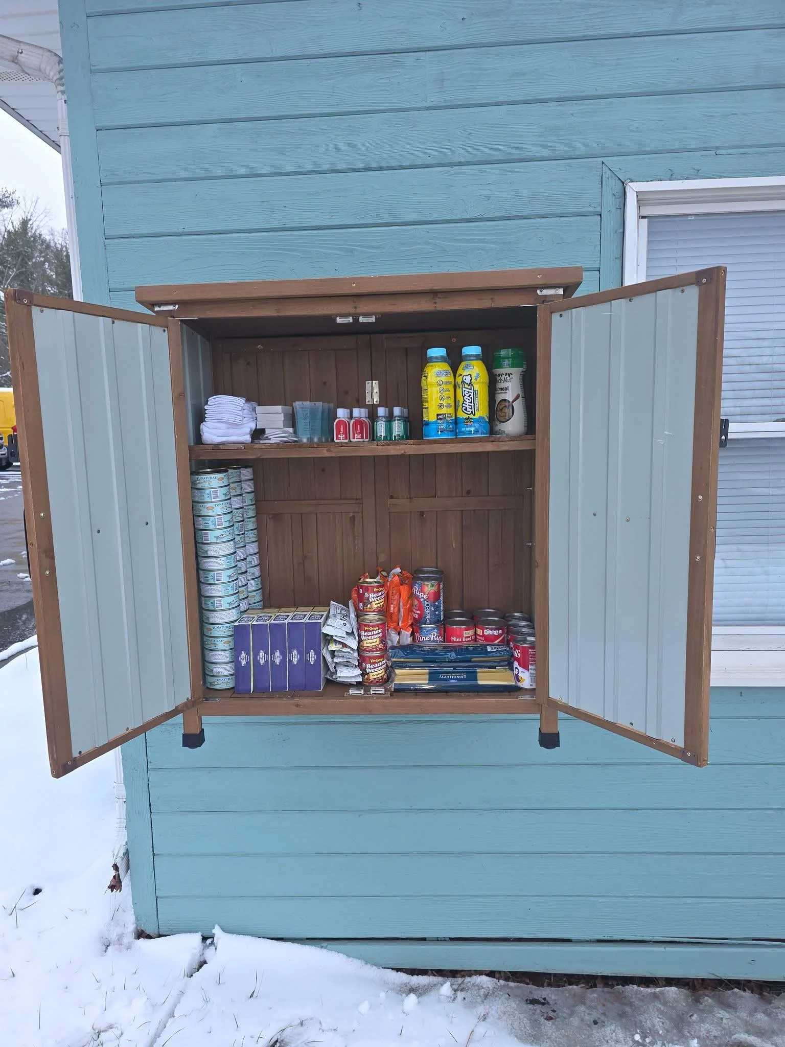 A wall-mounted, wooden outdoor food pantry is shown with both doors open, revealing shelves of food and household supplies inside. The pantry is attached to a teal-painted building exterior. Inside the cabinet are neatly arranged items including cann