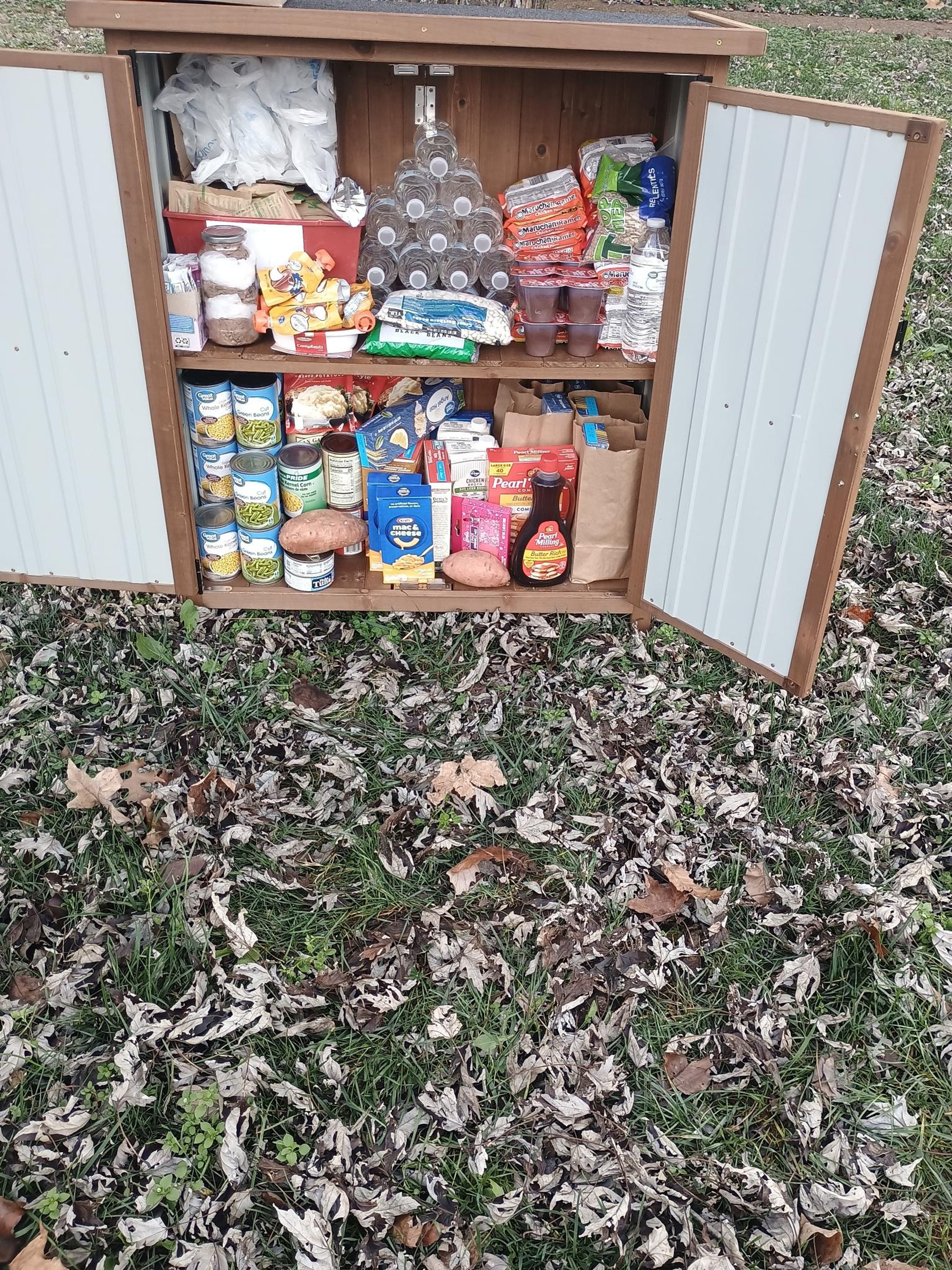 Open outdoor pantry on grass showing shelves with pasta, ramen noodles, canned meals, sauces, and shelf-stable milk.