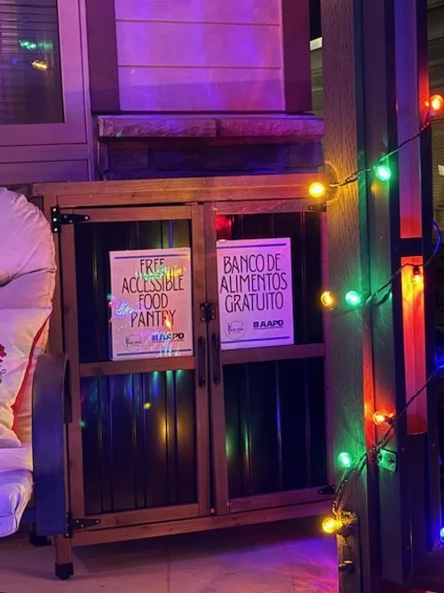 Closed outdoor food pantry at night, lit with colorful string lights, displaying bilingual signs for a free accessible food pantry.