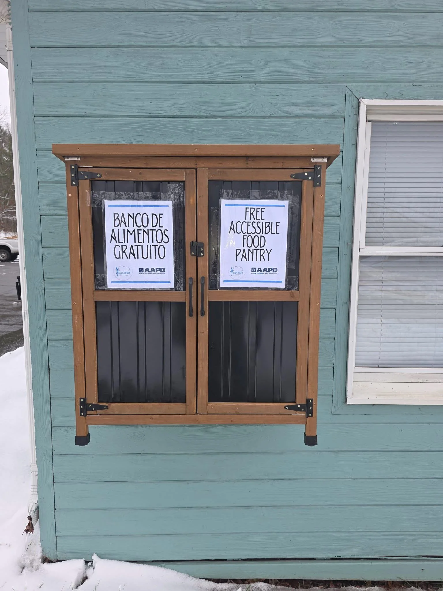 A wall-mounted, wooden outdoor food pantry is shown closed against a teal-painted building. The pantry has two front-opening doors with dark panels on the lower half and clear upper panels. Signs taped inside the doors read “Banco de Alimentos Gratui