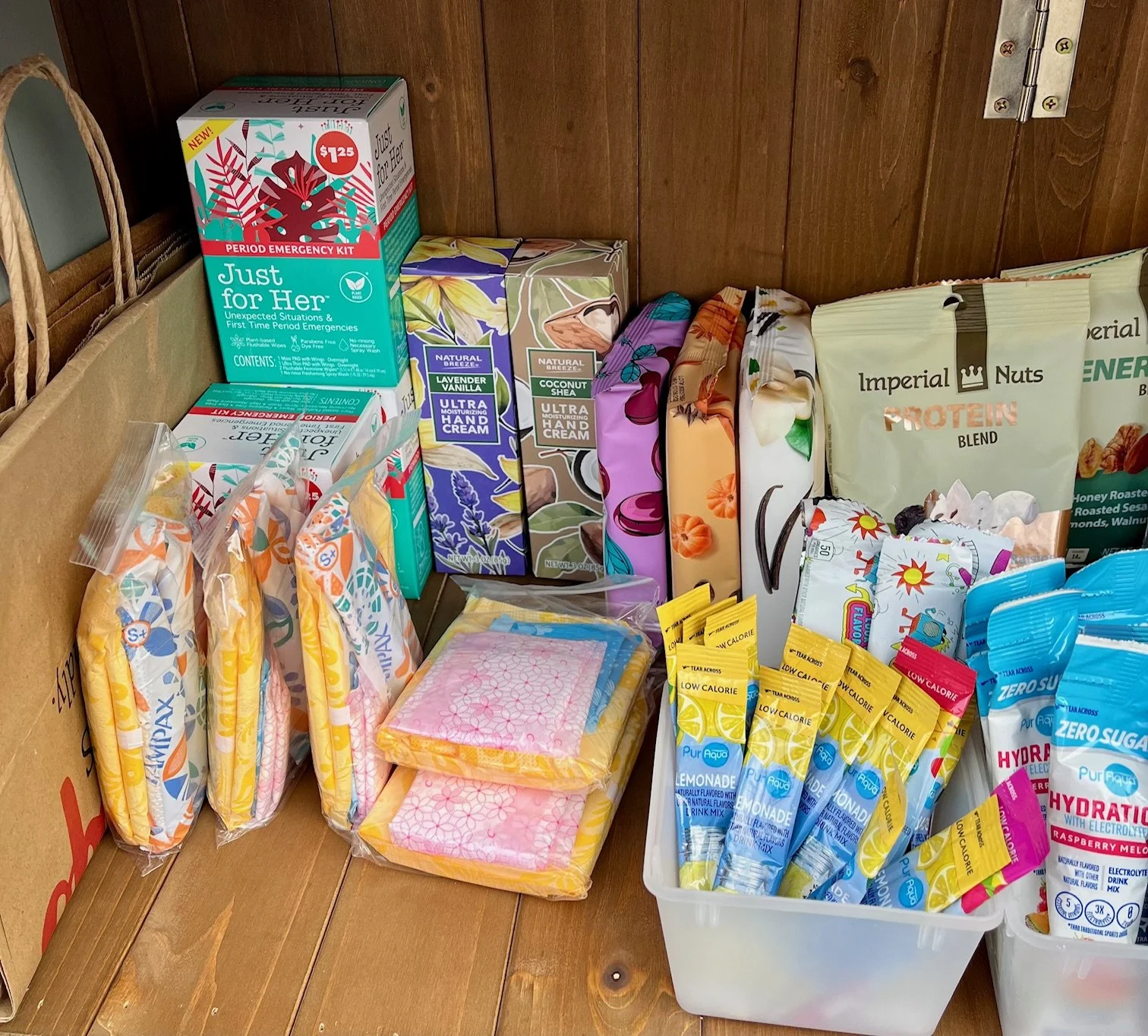 Close-up of pantry shelf with period supplies, hand cream, socks, reusable bags, hydration drink packets, and snack items.