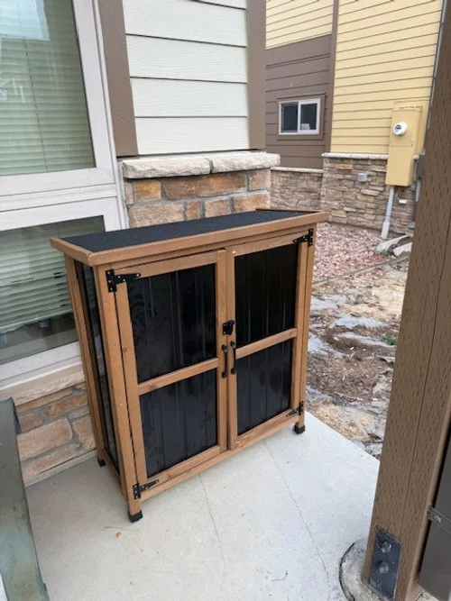 Closed wooden outdoor food pantry mounted on a concrete walkway beside a building, with dark-paneled doors and a slanted roof.