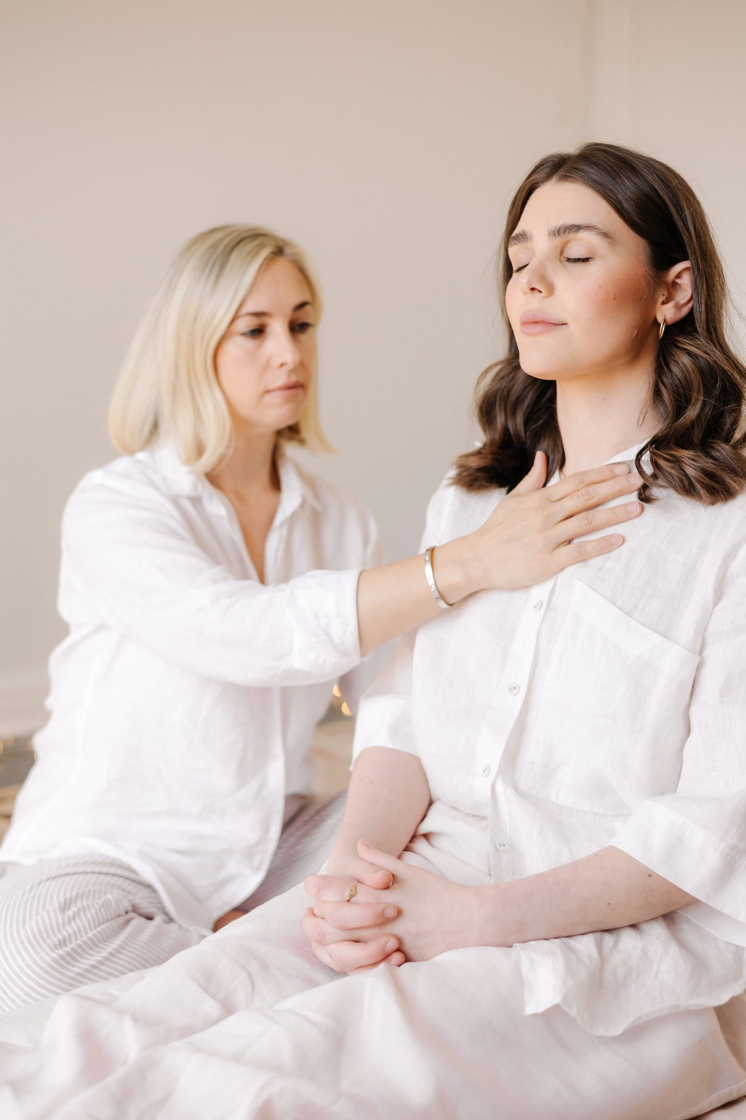 A woman with dark brown hair and light makeup has her eyes closed, gently touching her chest with her right hand while sitting on a bed. Another woman with blonde hair and light makeup stands behind her, placing her left hand on the woman's shoulder and her right hand on her chest. Both women are wearing white shirts in a softly lit room with a neutral wall background.