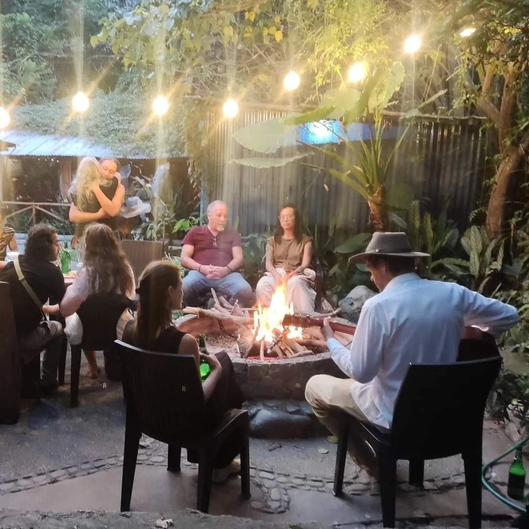 People gathered around a fire pit in a backyard at night, some sitting and some standing, with warm lighting overhead and lush greenery in the background.