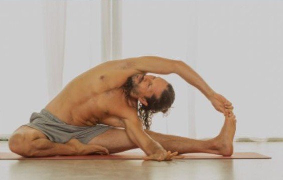 Older man practicing seated forward bend yoga stretch on mat in a bright room.