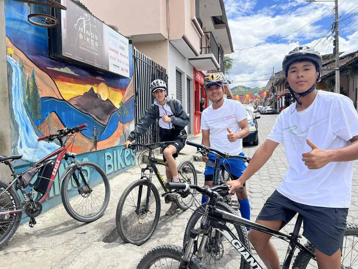 Three young men in cycling gear posing with their bikes on a street with colorful murals and houses, giving thumbs up.