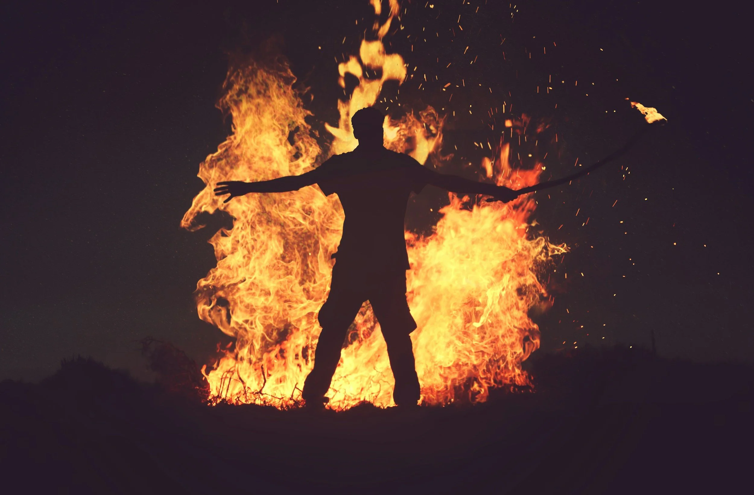 Silhouette of a person standing with arms outstretched in front of a large fire at night.