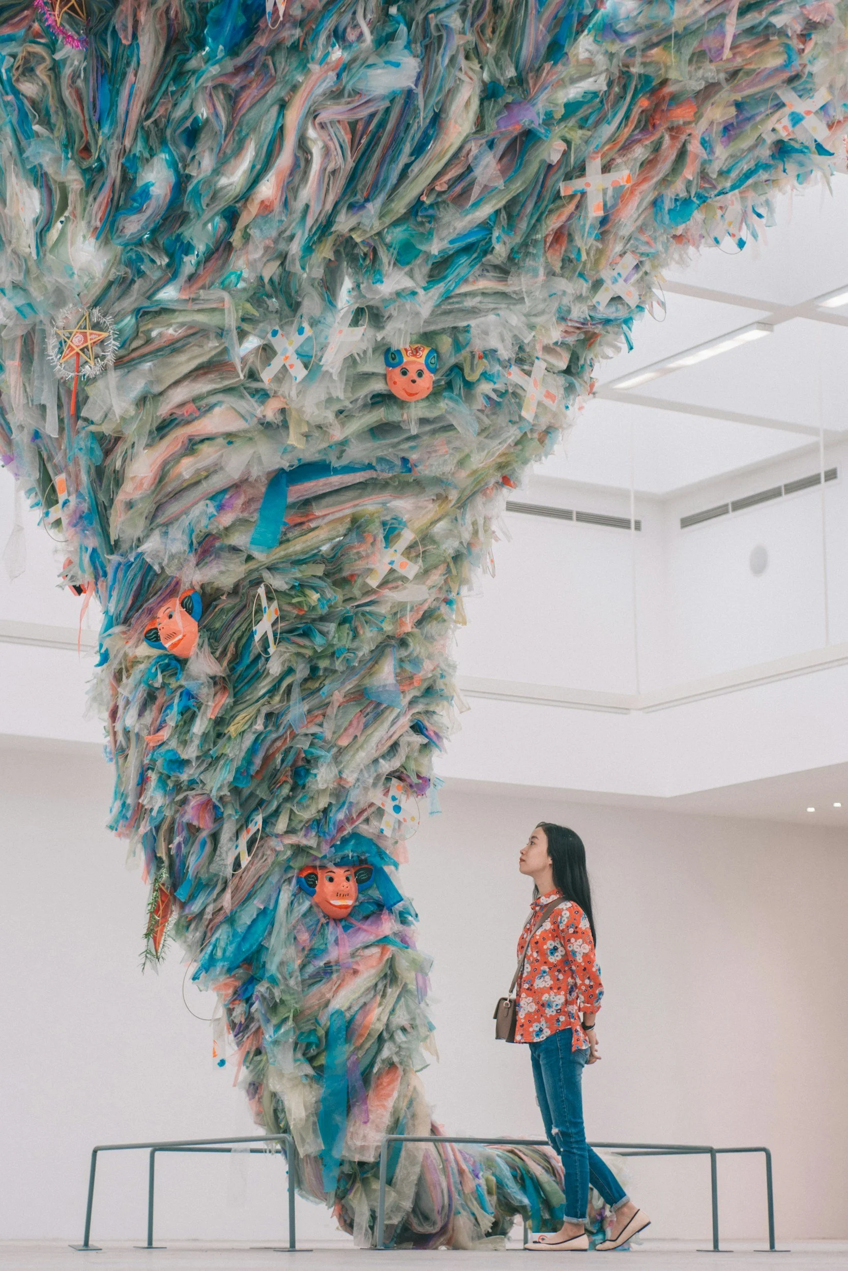 A woman stands beside an art installation made of colorful, layered fabric and features several masks with expressive faces and animal designs. The installation curves from the ceiling to the floor in a flowing form.
