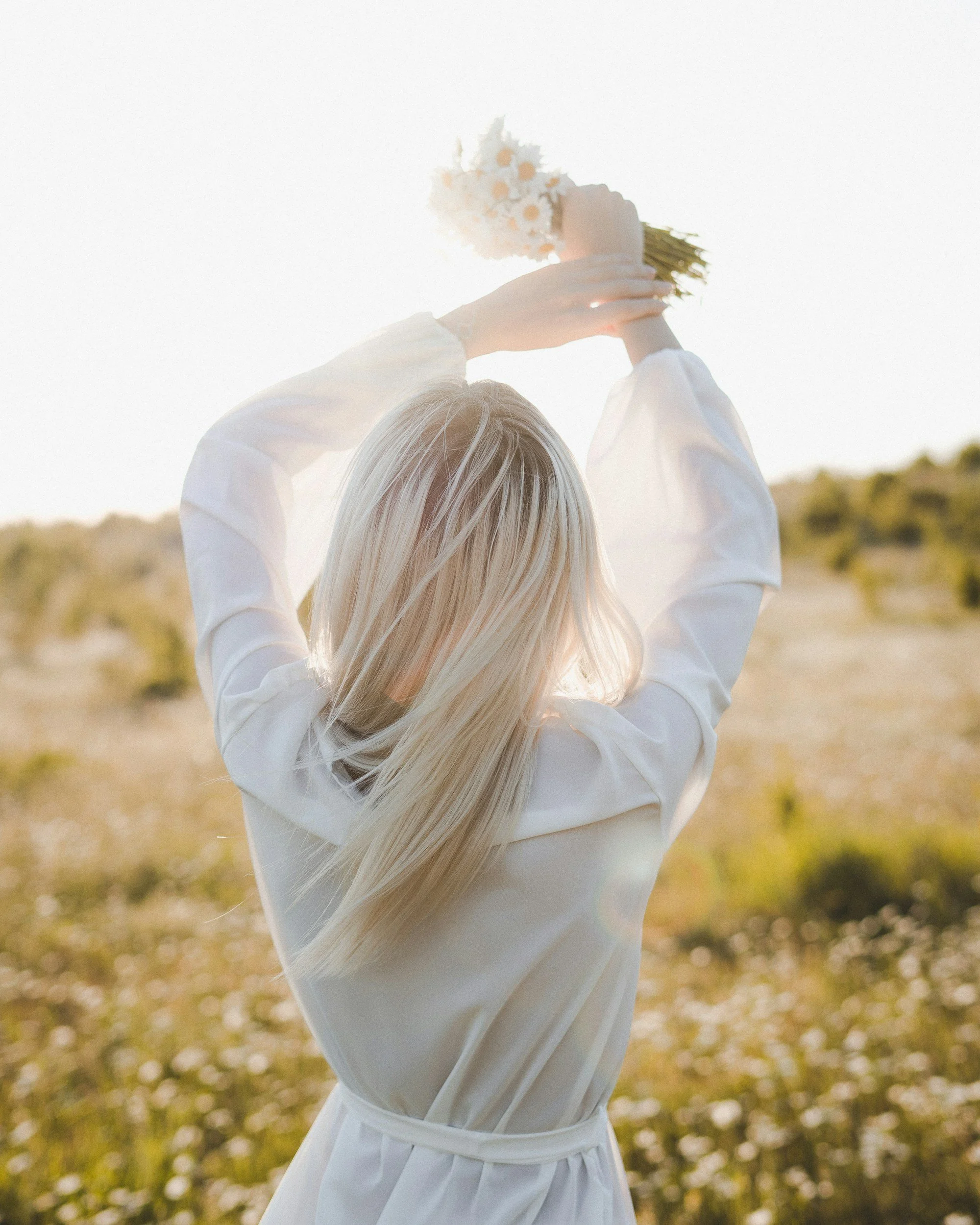 Person with long blonde hair holding a bouquet of daisies against a bright sky in a field.
