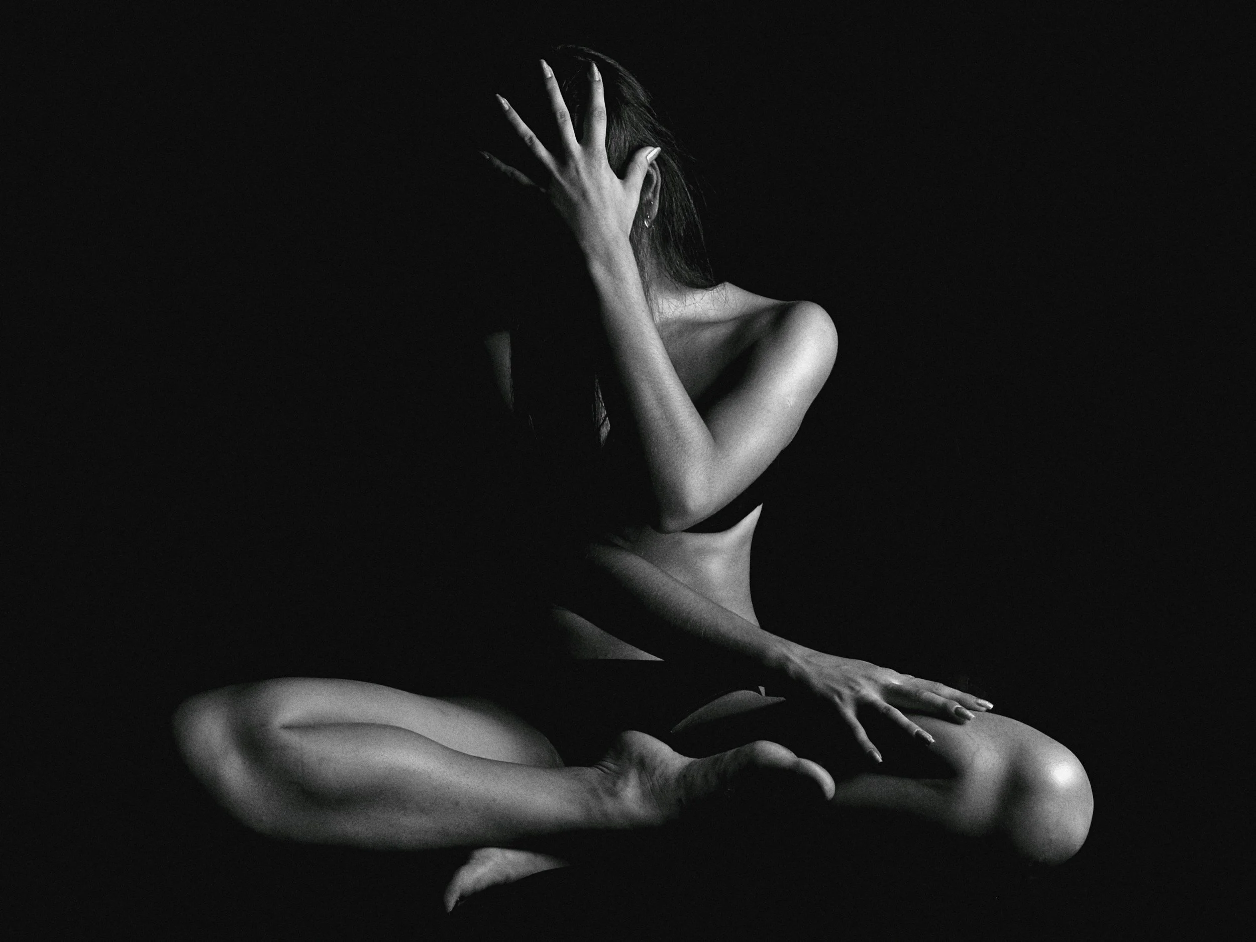 Black and white photograph of a nude woman kneeling and covering her face with her hand, with another hand resting on her leg, against a dark background.