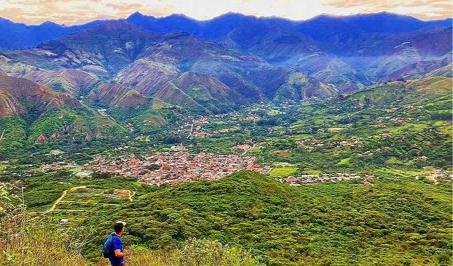 A person standing on a hillside overlooking a lush green valley with a town, surrounded by mountains under a cloudy sky.