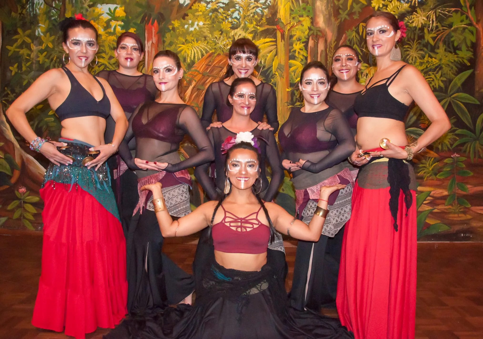 Group of nine women in traditional and dance costumes with painted faces, posing together in a lush, jungle-like setting with dense green foliage in the background.
