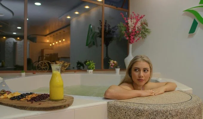 A young woman relaxing in a spa or hot tub with a pitcher of yellow juice, a wooden tray with berries, and pink flowers in a large white vase nearby.