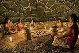 Seven people sitting in a circle inside a tent, participating in a group meditation or prayer around a small fire with a candle in the center.