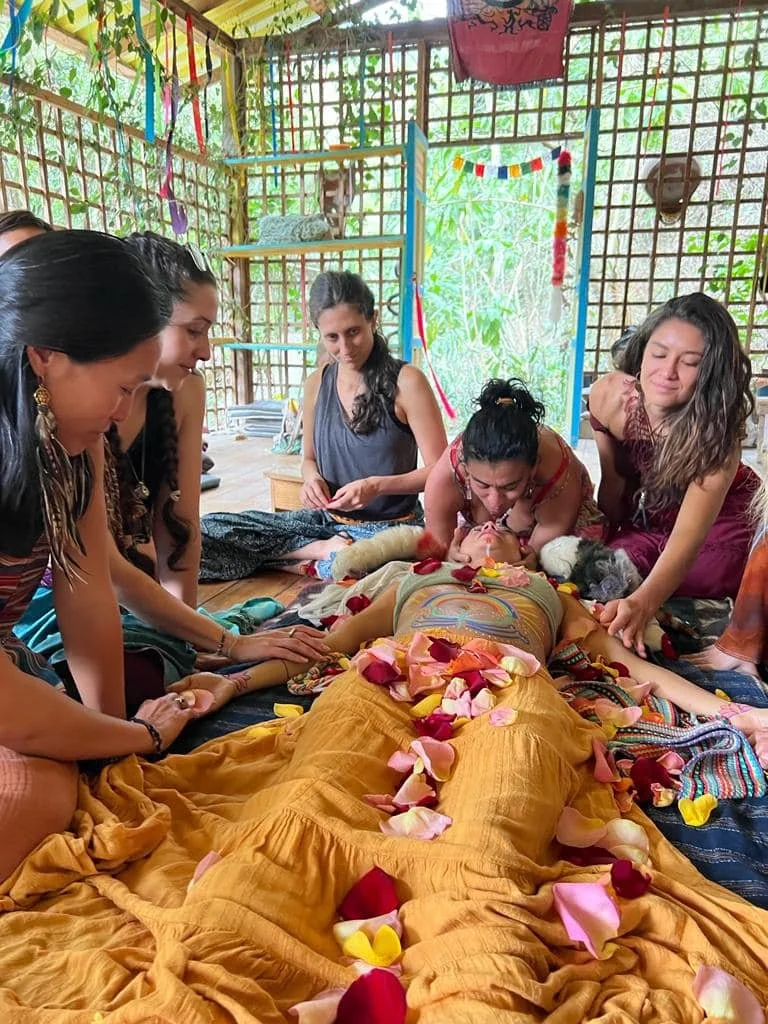 Group of women performing a ritual or ceremony around a person lying on a bed decorated with rose petals, in an open, lush outdoor setting.