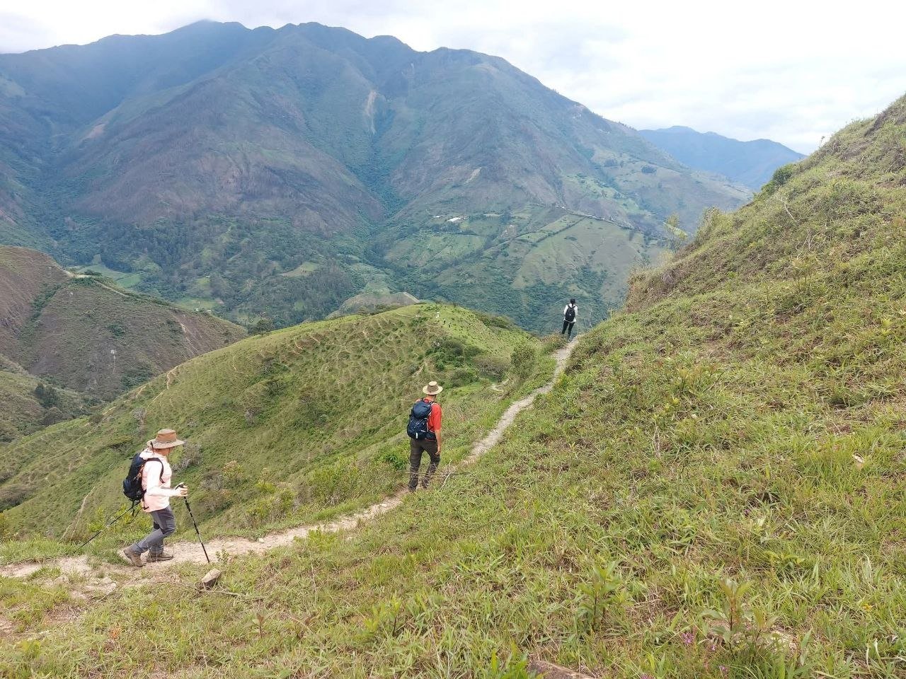 Three hikers with backpacks walking along a mountain trail surrounded by green hills and mountains in the distance.