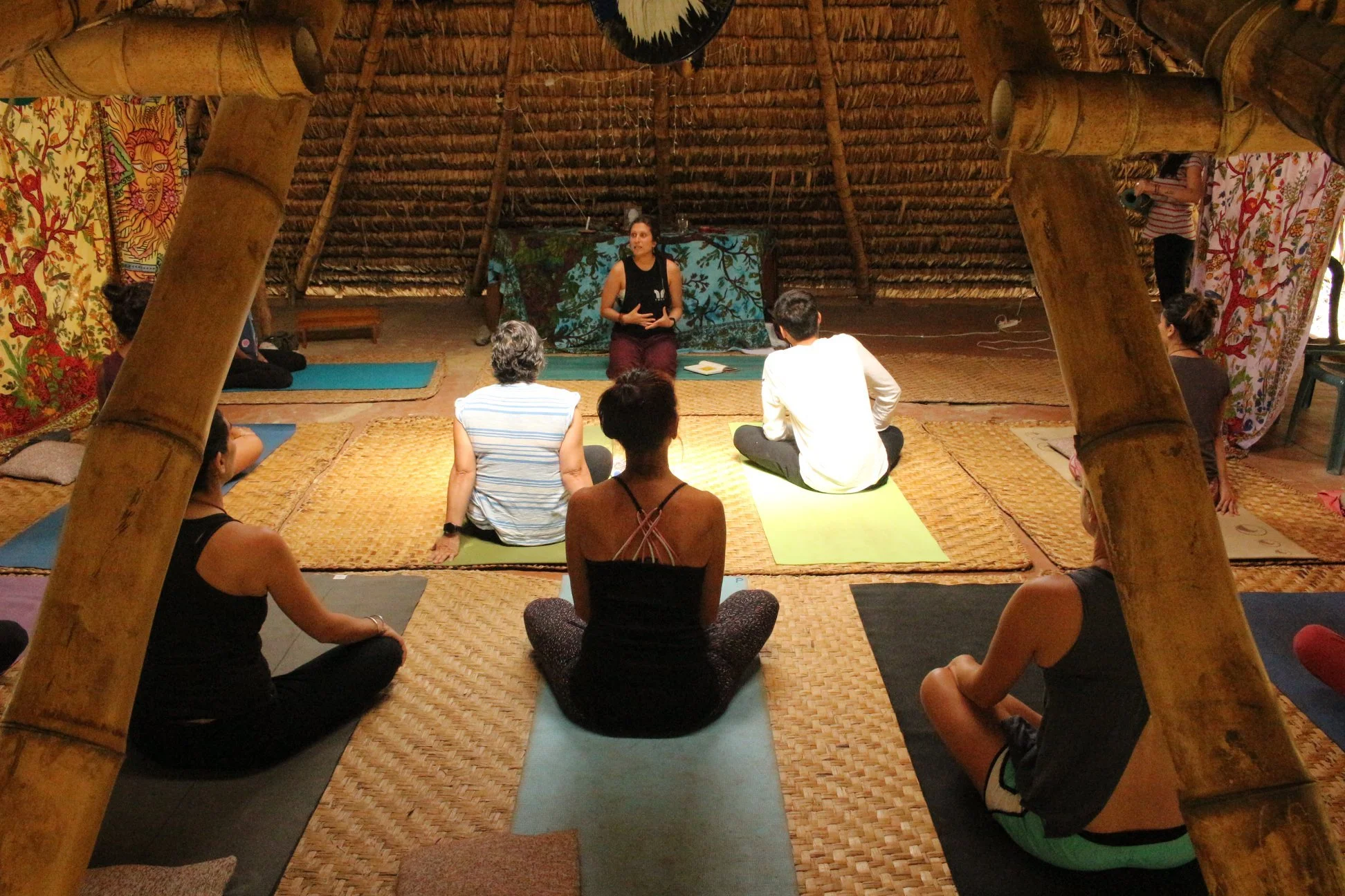 A woman leading a yoga class in a rustic, thatched-roof room with tropical decor. Participants are seated on yoga mats, facing her, engaged in a guided meditation or breathing exercise.