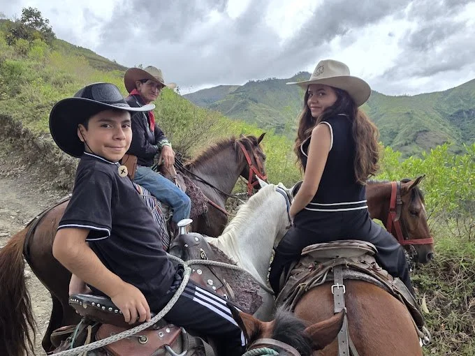 Two children and an older woman horseback riding on a trail in a mountainous, green landscape, all wearing cowboy hats.