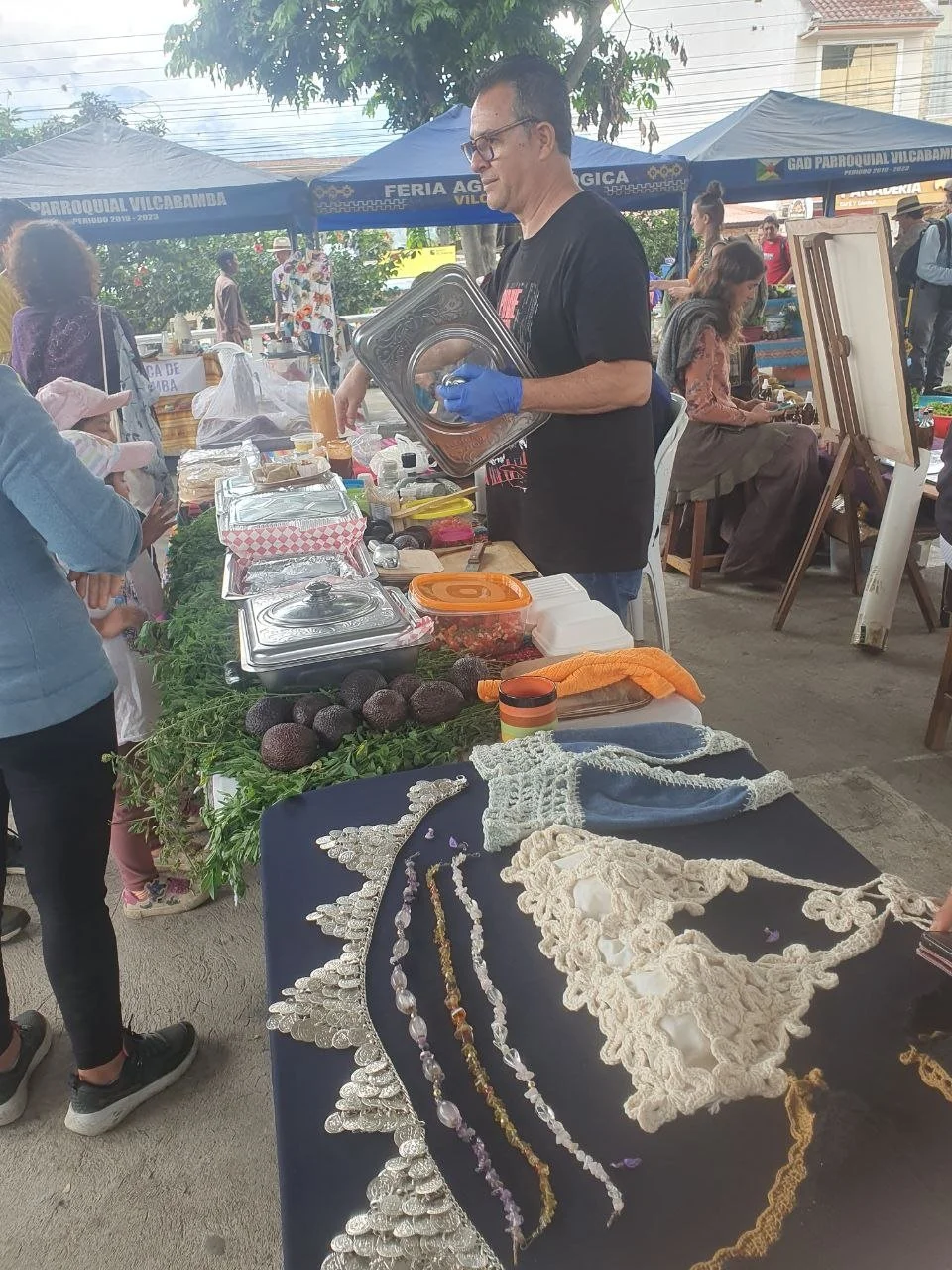 A man wearing glasses and gloves, standing at a market stall, arranging metal trays with food. The stall also displays avocados, a glass of juice, and various jewelry on a dark cloth. There are other market vendors and customers in the background, with tents and trees in an outdoor setting.