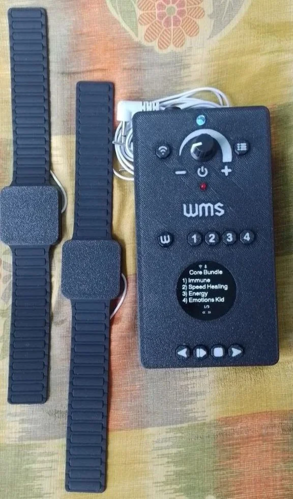 A remote control device labeled 'WMS' with various buttons and a black box attached to two adjustable straps with textured surfaces, all on a multicolored fabric surface.